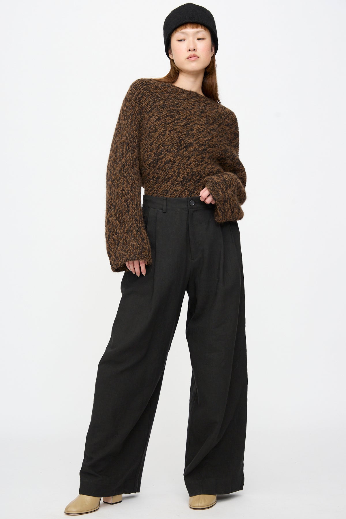 A person wearing a brown chunky knit sweater, Lauren Manoogian Soft Trouser in Black, a black hat, and tan boots.