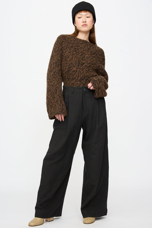 A person wearing a brown chunky knit sweater, Lauren Manoogian Soft Trouser in Black, a black hat, and tan boots.