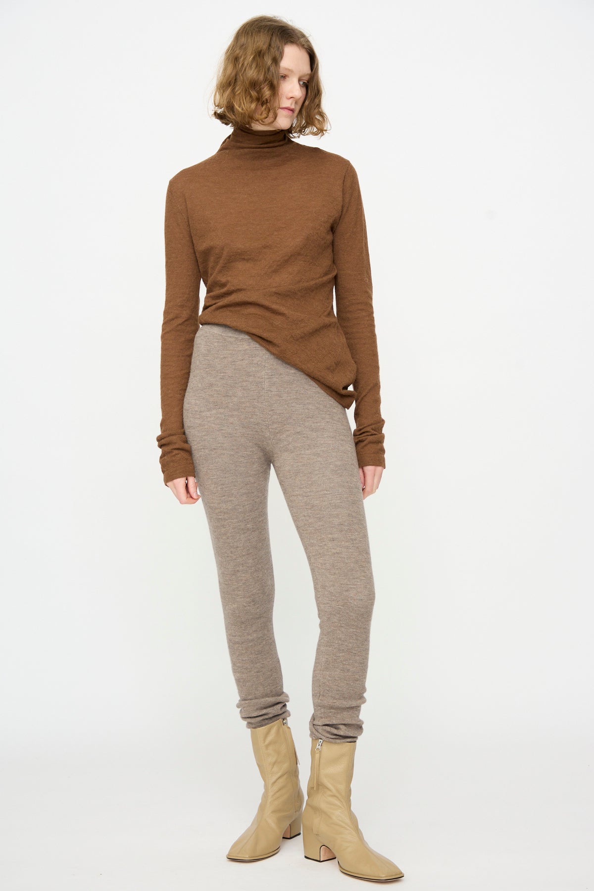 A person wearing a brown turtleneck, the Lauren Manoogian Stirrup Legging in Grey Melange, and beige ankle boots stands against a plain white background.