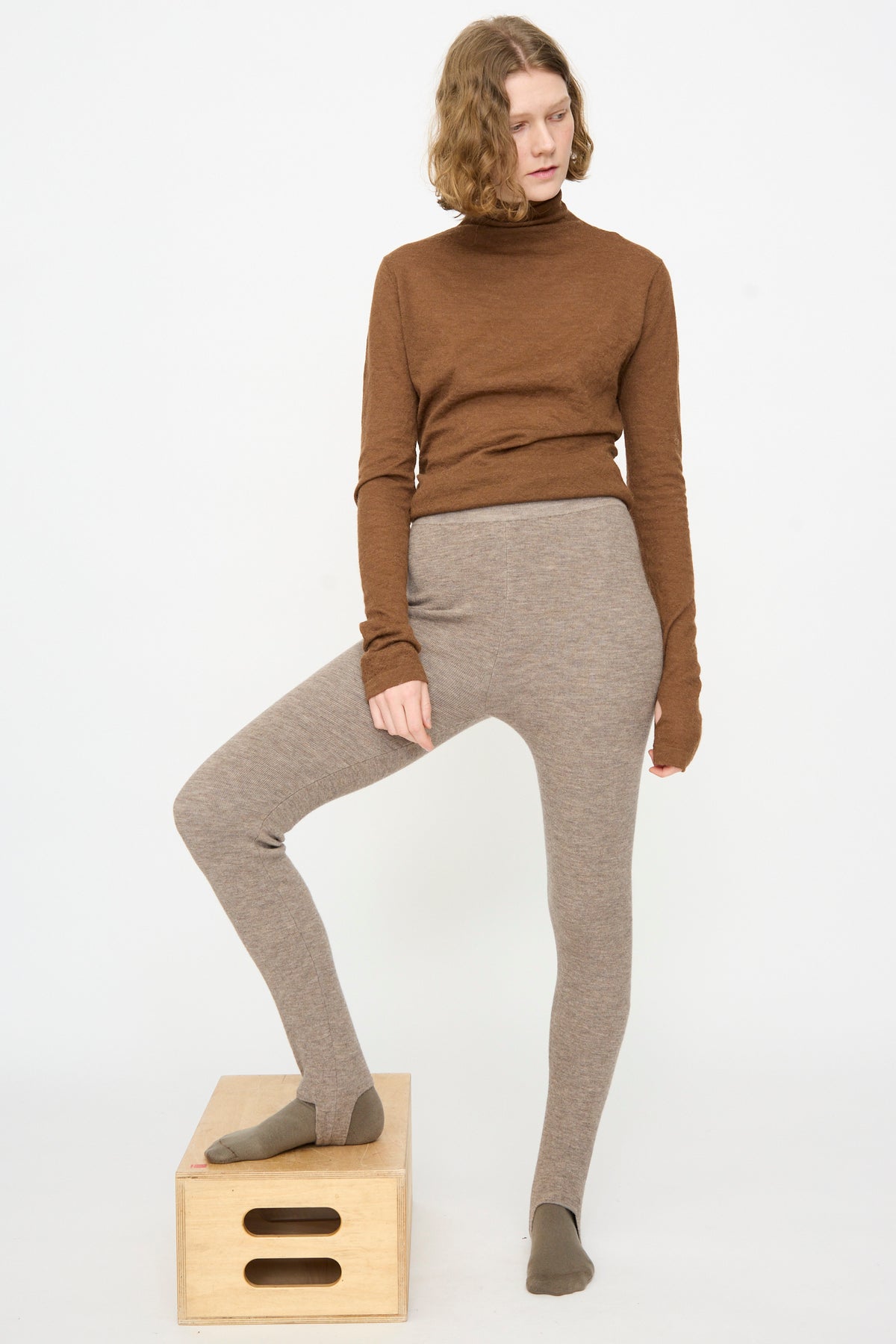 A person wearing a brown turtleneck, Lauren Manoogian Stirrup Legging in Grey Melange, and brown socks stands with one foot on a wooden box against a plain white background.