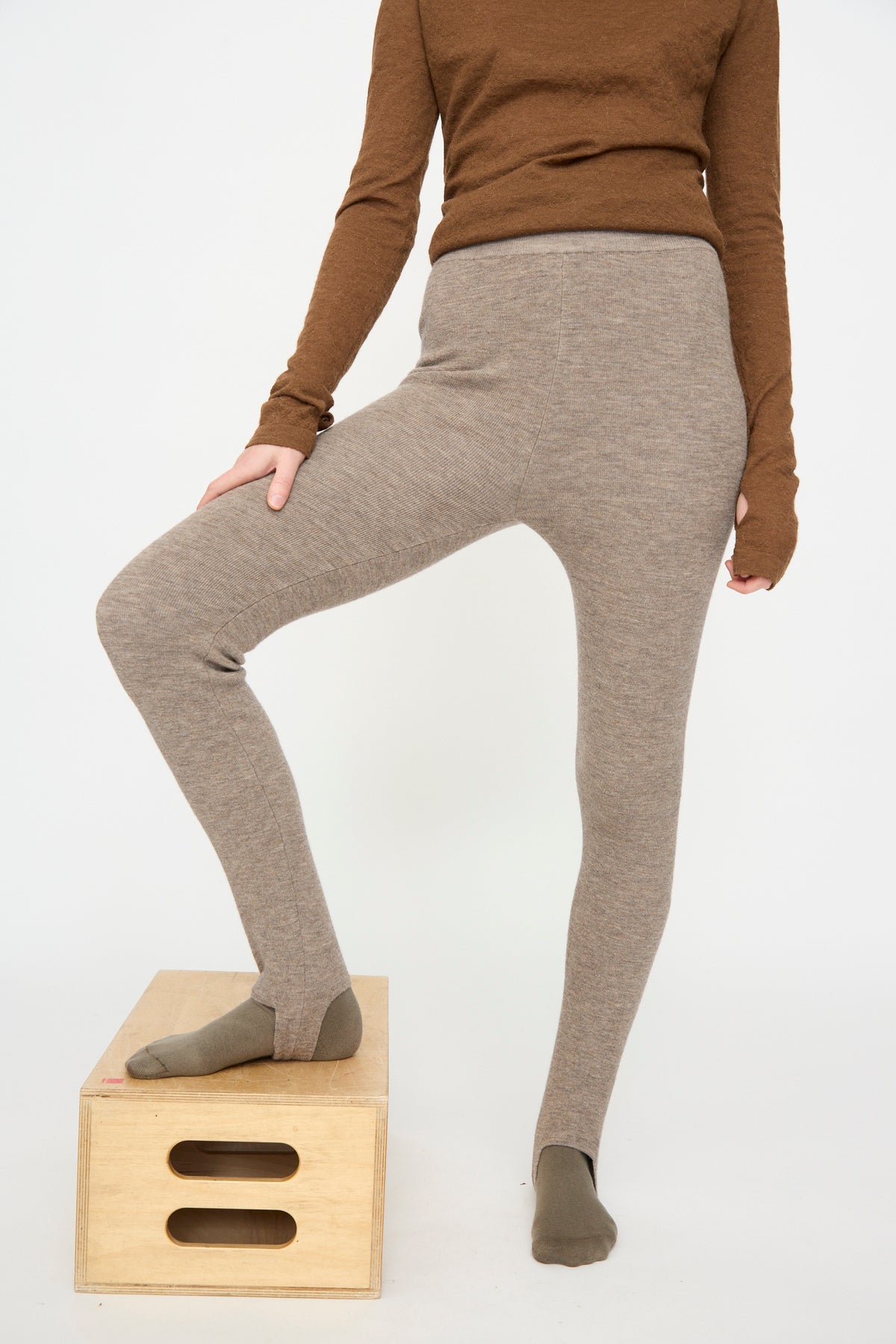 A person wearing a brown long-sleeve top and the Lauren Manoogian Stirrup Legging in Grey Melange stands with one foot on a wooden box against a plain white background.