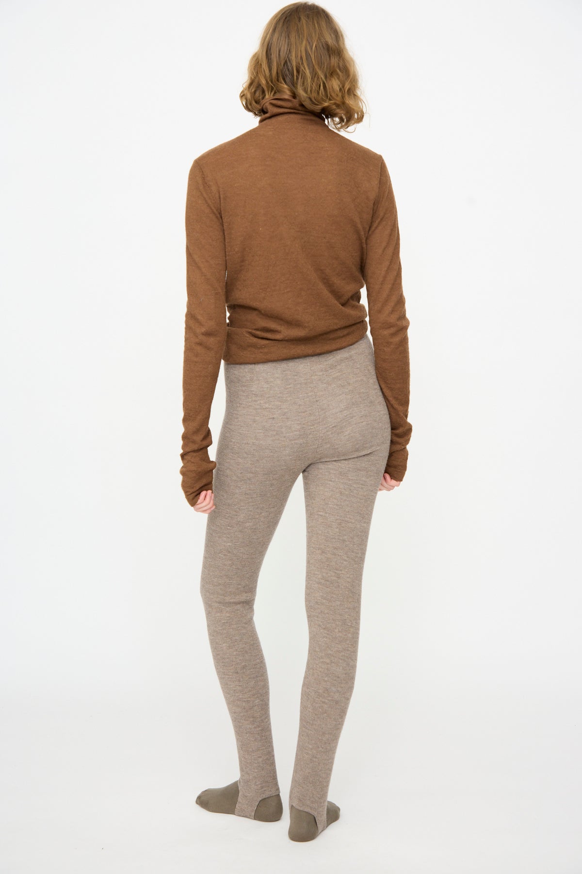 A person stands facing away from the camera, wearing a brown long-sleeve top and Lauren Manoogian’s Stirrup Legging in Grey Melange. The background is plain white.