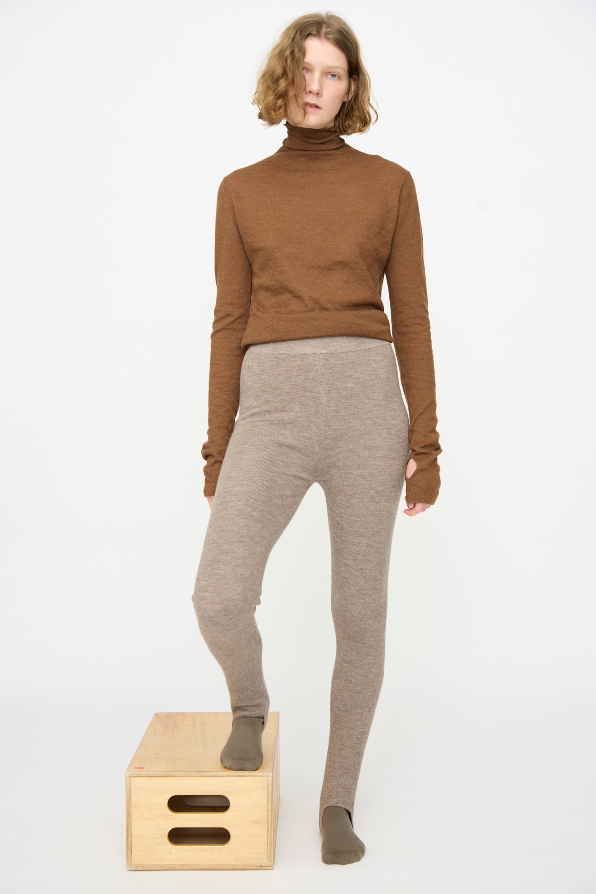 A person wearing a brown turtleneck top and the Lauren Manoogian Stirrup Legging in Grey Melange stands with one foot on a wooden box against a plain white background.