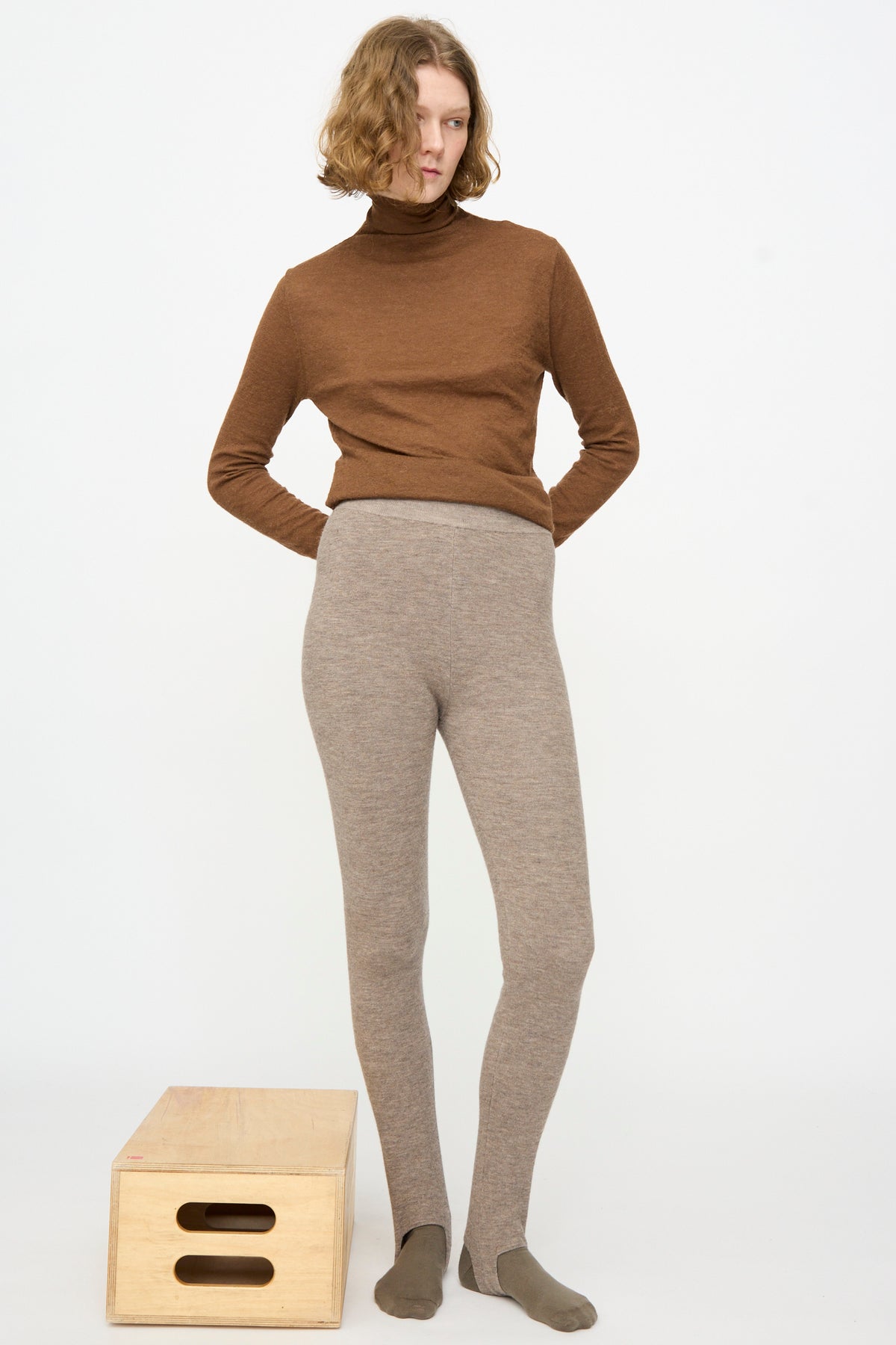 A person stands next to a wooden box, wearing a brown turtleneck sweater and Lauren Manoogian’s Stirrup Legging in Grey Melange, against a plain white background.