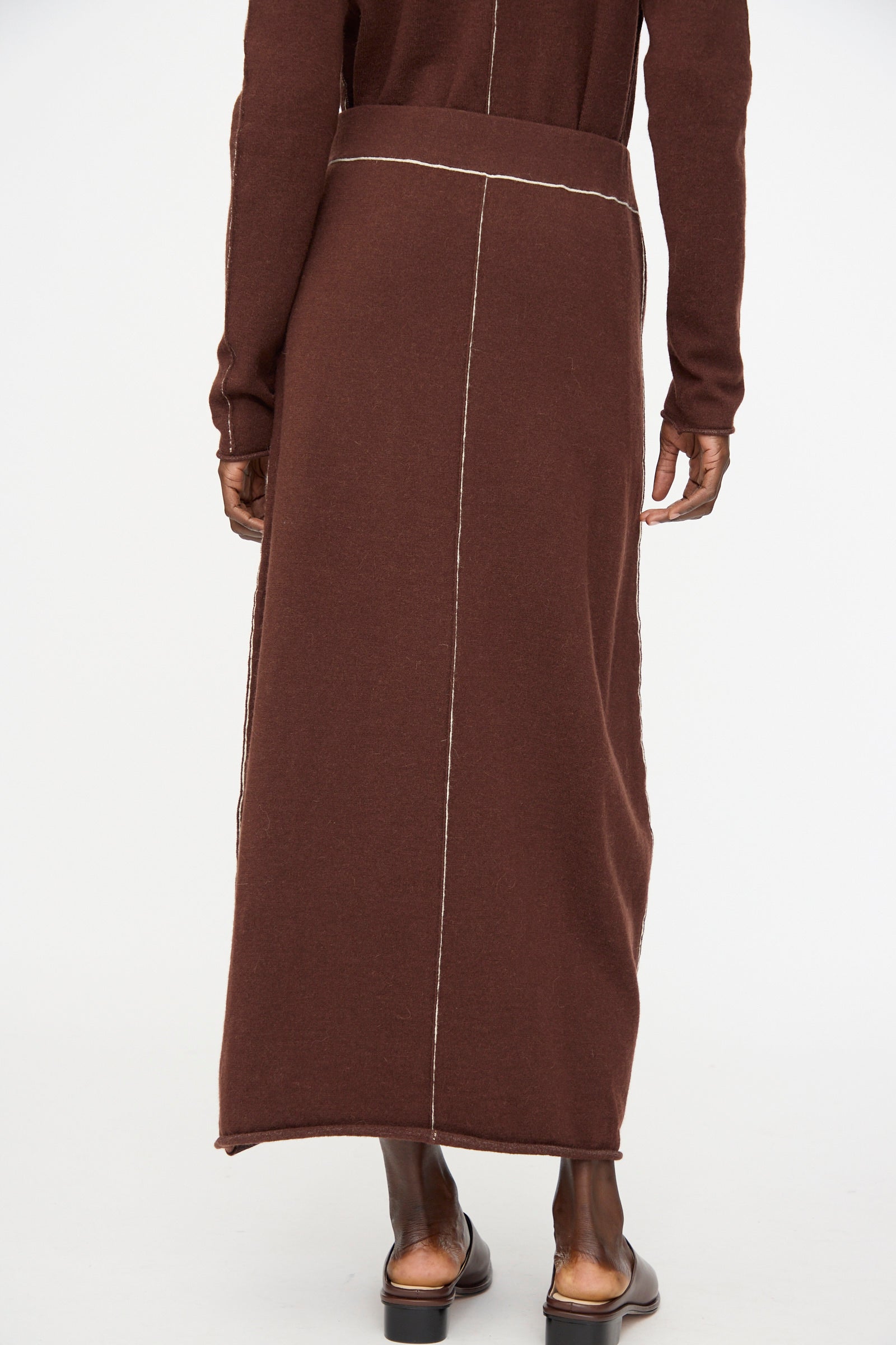 A person stands with hands by their sides, wearing the Lauren Manoogian Stitch Skirt in Merlot paired with flat, open-back shoes. Visible white stitching runs down the back of the skirt.