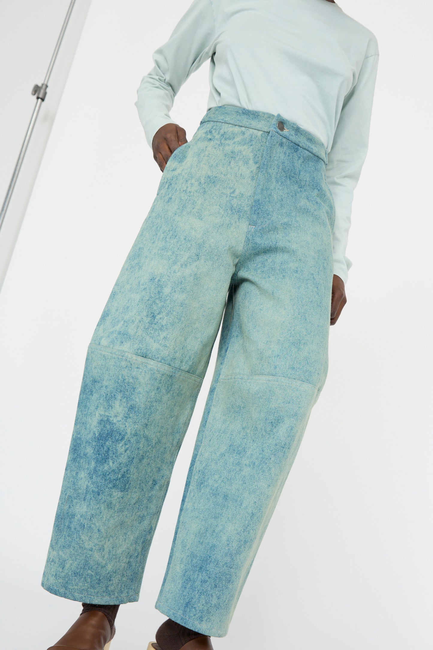 A person wearing Lauren Manoogian's Structure Jean in Acid, a high-waisted, wide-legged, relaxed fit light blue denim and a light green long-sleeve shirt stands against a plain white background.