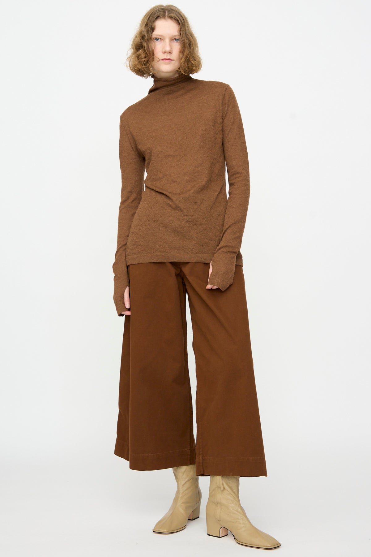 A person wearing a brown turtleneck, Lauren Manoogian Sweep Pants in Rust—relaxed fit cotton pants made in Peru—and beige ankle boots stands against a plain white background.