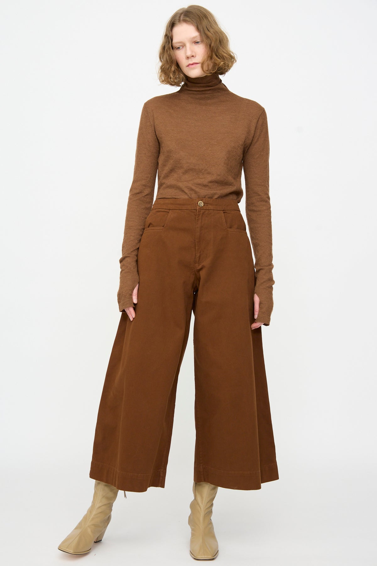 A person stands against a white background, wearing a brown turtleneck sweater, Lauren Manoogian Sweep Pants in Rust—wide-legged relaxed fit cotton pants made in Peru—and beige boots.