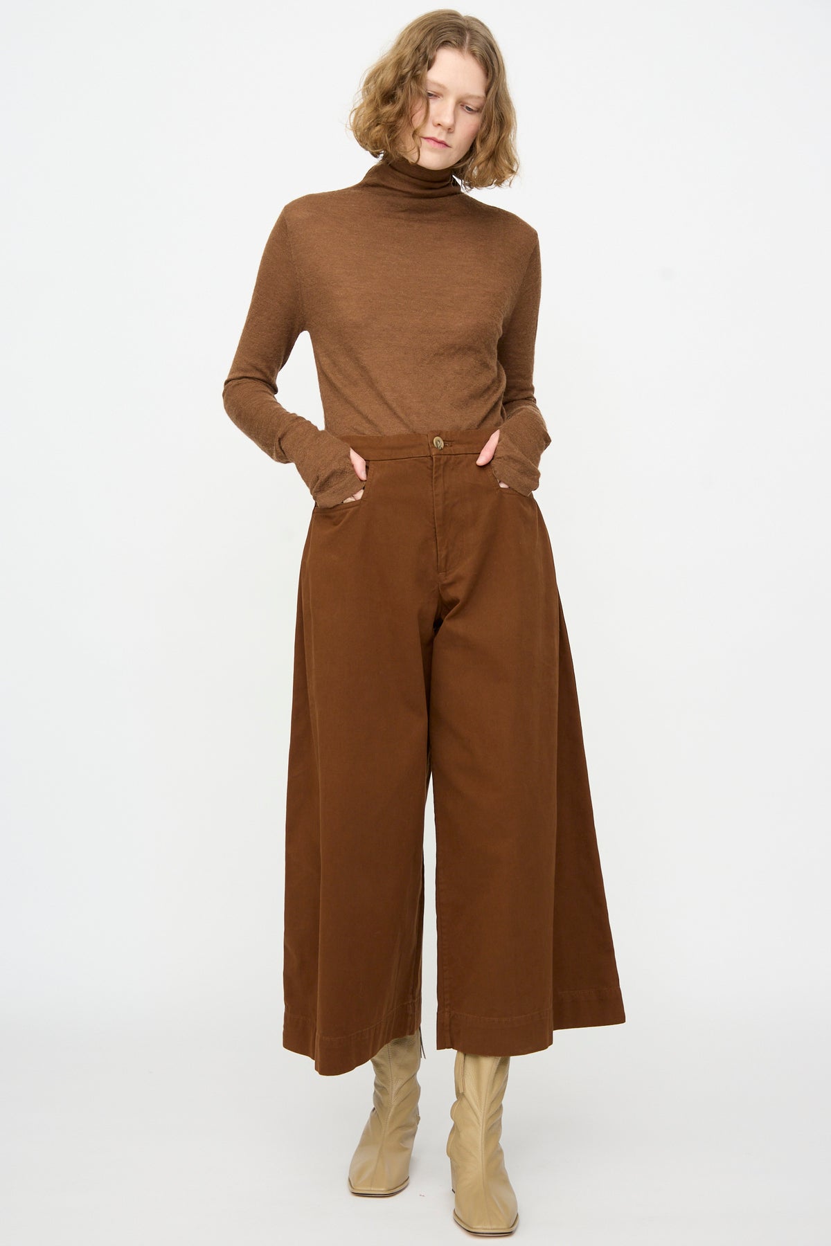 A person stands against a plain background wearing a brown turtleneck sweater, Lauren Manoogian’s Sweep Pants in Rust, and beige boots.