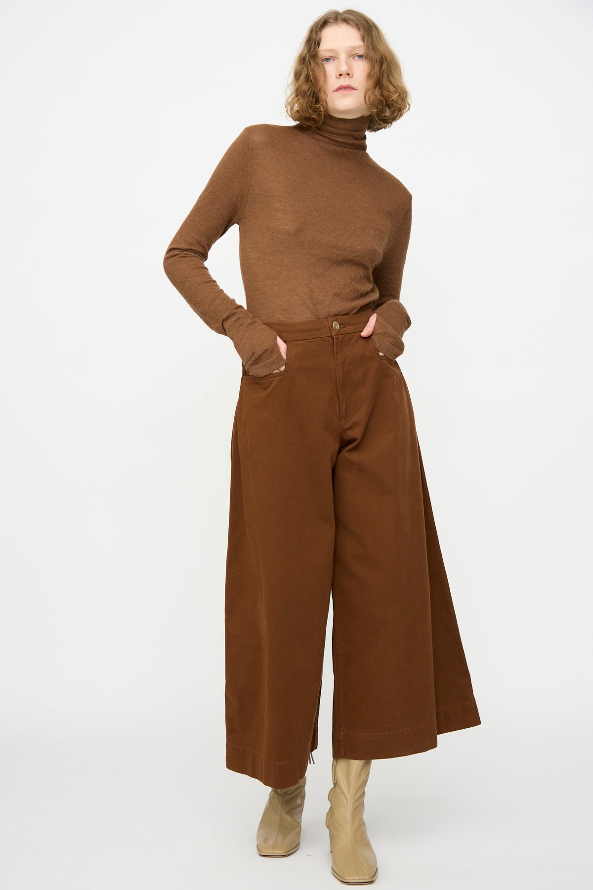 A person stands against a plain background, wearing a brown turtleneck sweater and Lauren Manoogian’s Sweep Pants in Rust—wide-legged cotton pants with a relaxed fit—paired with beige boots. Their hands are in their pockets and their hair falls in loose waves.