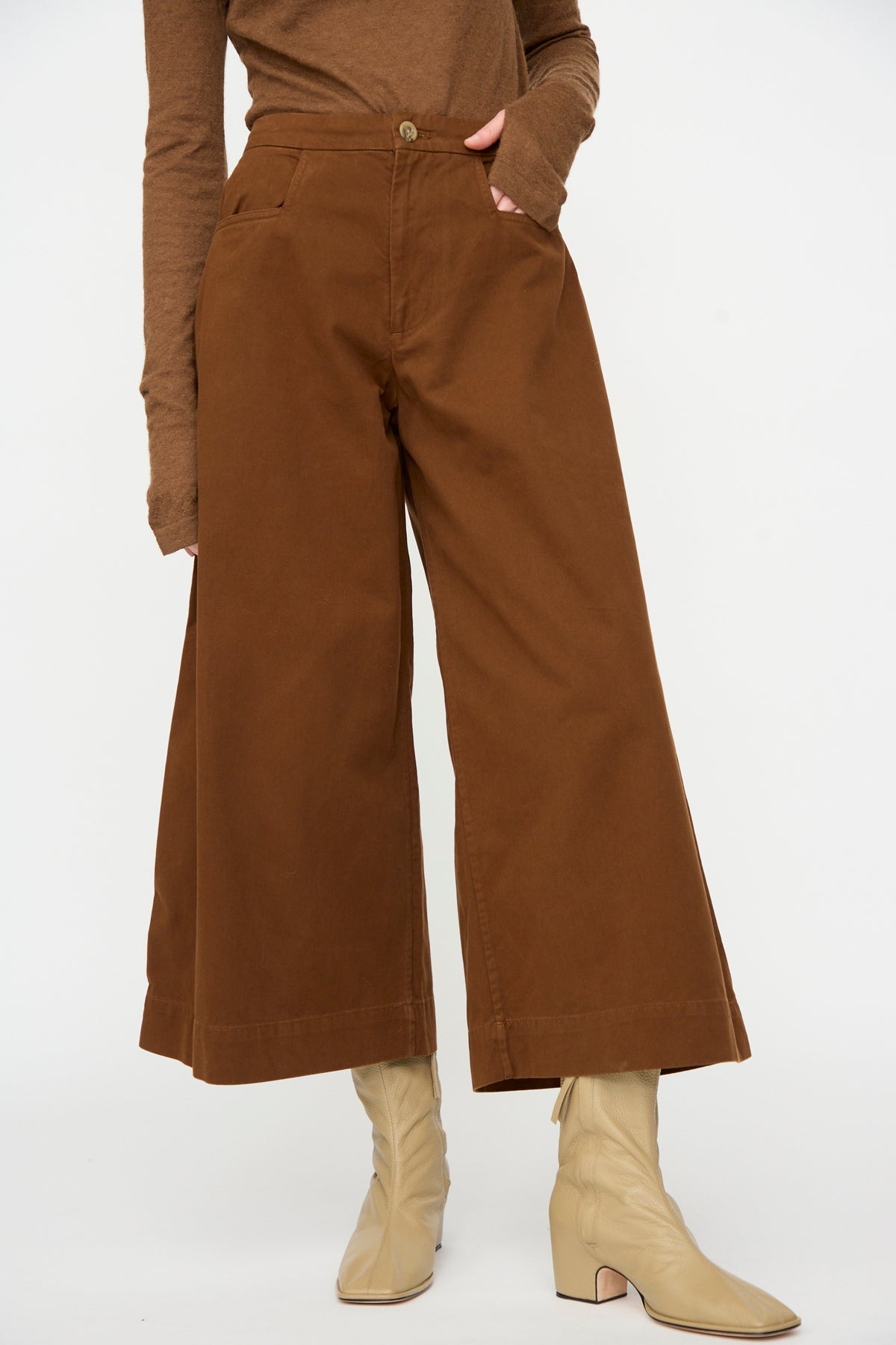 A person wearing Lauren Manoogian’s Sweep Pants in Rust, paired with a matching brown long-sleeve top and beige ankle boots, stands against a plain white background.
