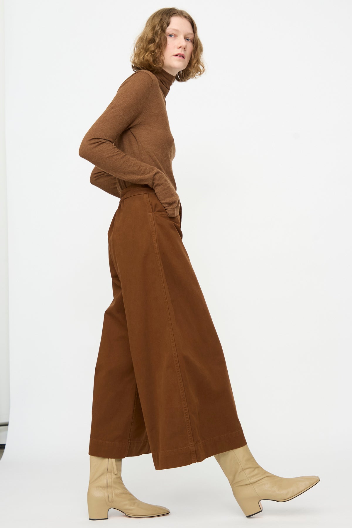 A person wearing the Lauren Manoogian Sweep Pants in Rust, paired with a brown turtleneck and light beige boots, stands against a plain white background.