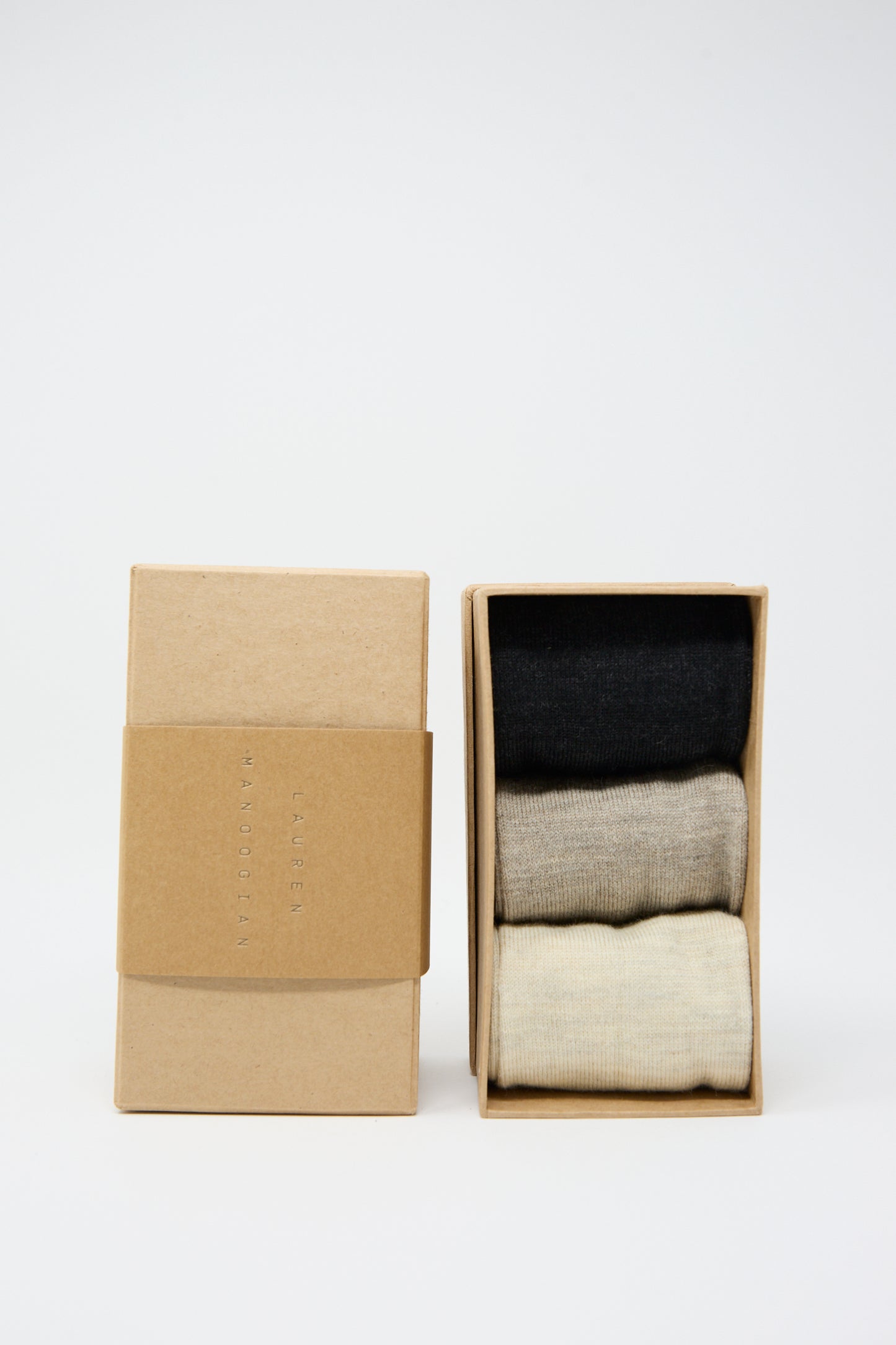 Two cardboard boxes, one open and one closed. The open box contains a pack of three neatly folded Tall Socks in Mix by Lauren Manoogian in black, gray, and beige colors. The boxes have simple, minimalistic brown packaging.