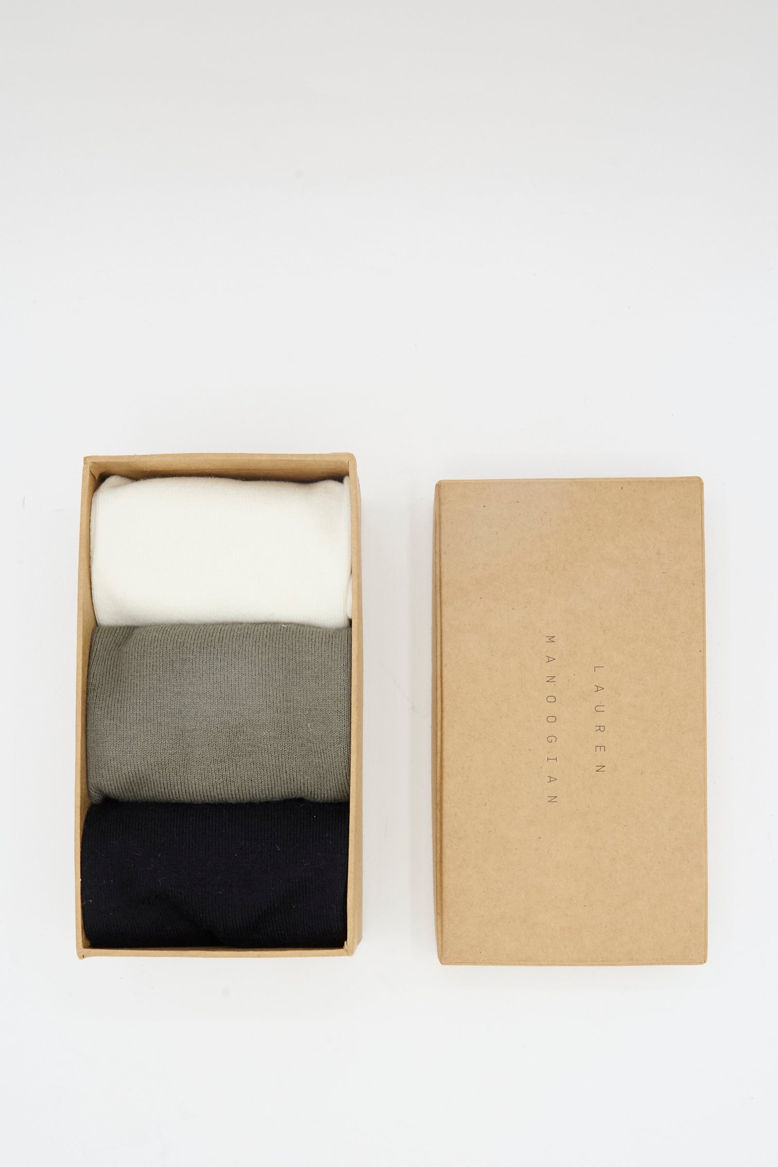A cardboard box labeled "Lauren Manoogian" holds three neatly folded Tall Socks in Mix—white, gray, and black alpaca wool.