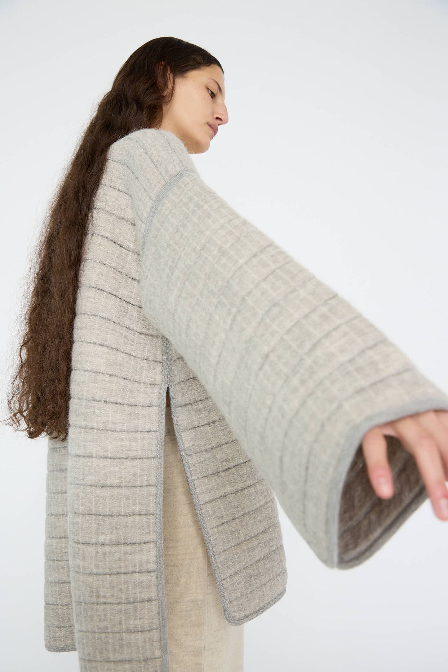 A person with long hair is wearing a Tami Pullover in Grey Melange by Lauren Manoogian, stretching an arm out against a plain background.