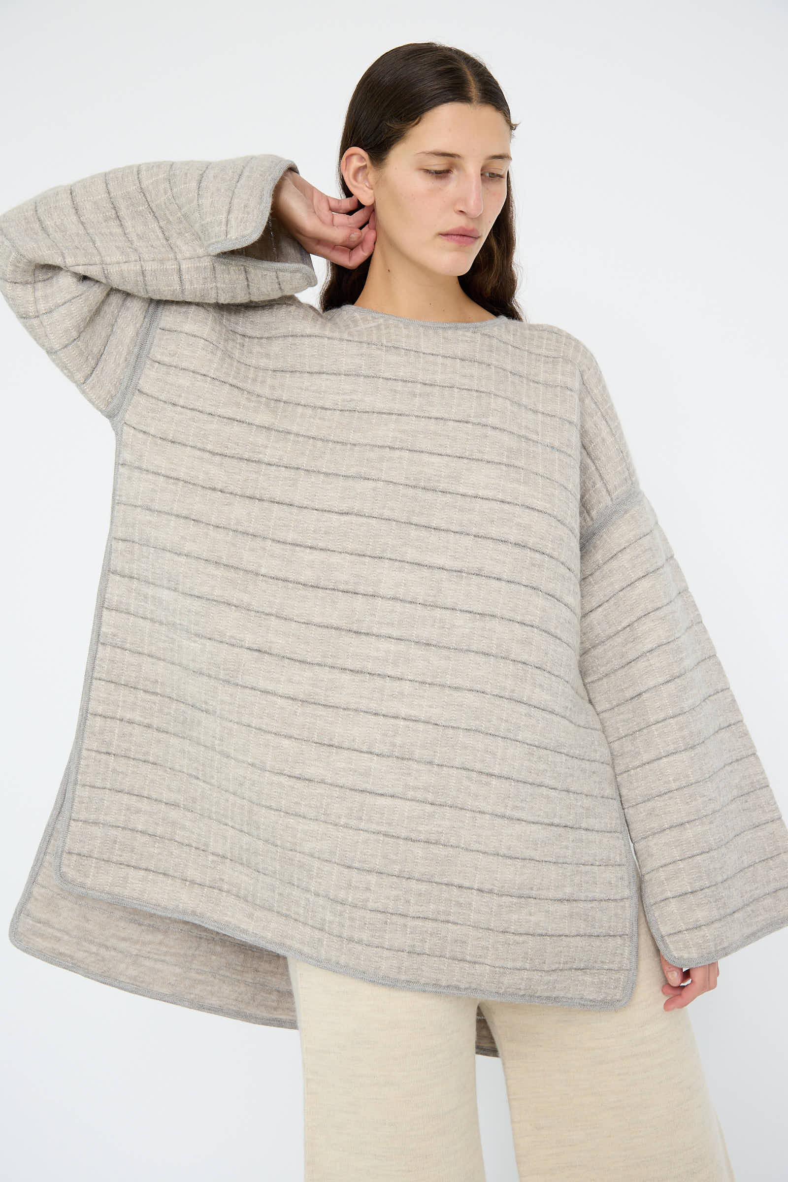 A person wearing the Tami Pullover in Grey Melange by Lauren Manoogian, which is an oversized, striped, long-sleeve sweater made from quilted knit alpaca, paired with light beige pants for a relaxed fit, against a plain white background.