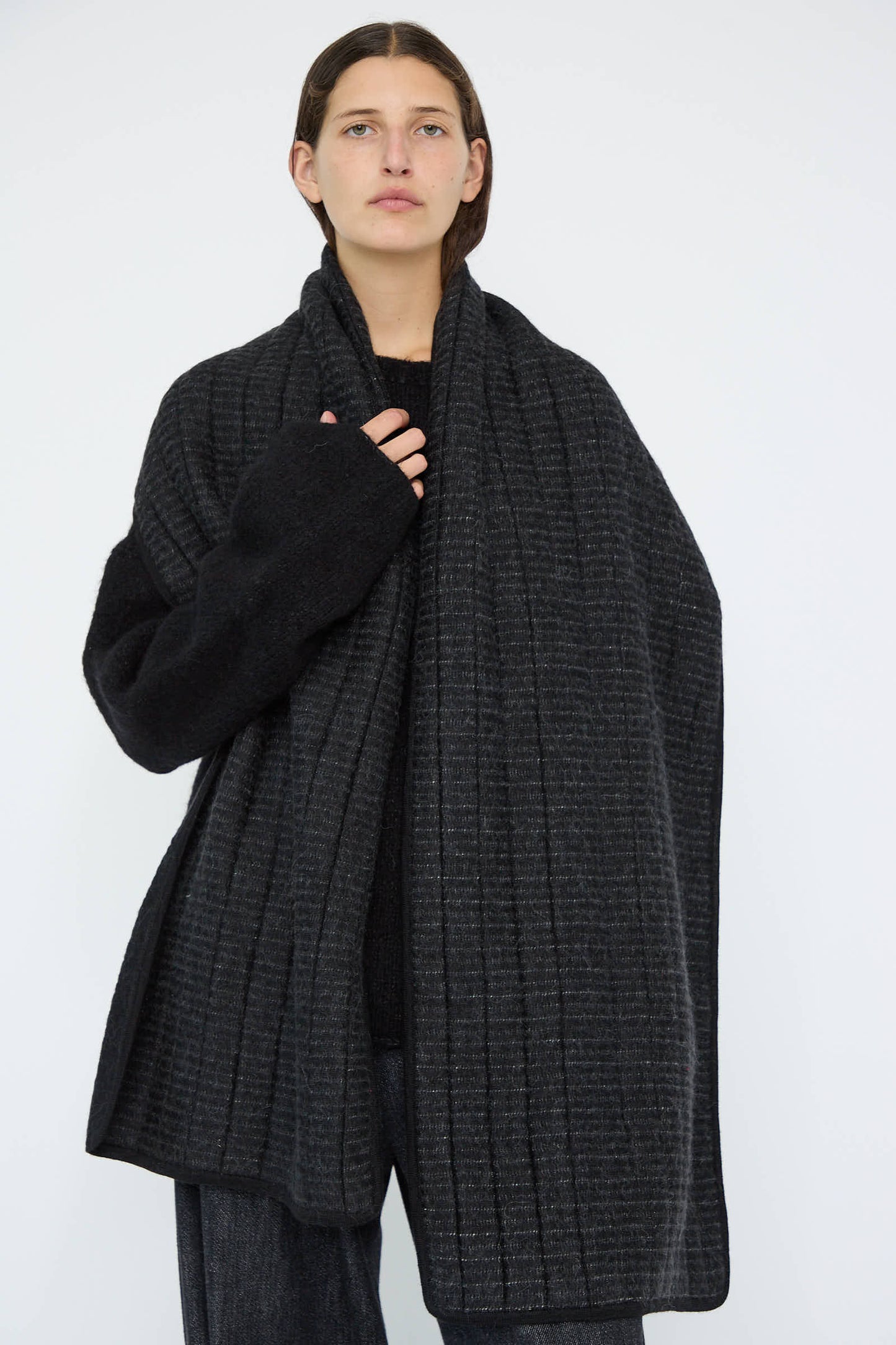 A person is seen wearing the Lauren Manoogian Tami Wrap in Black, a beautifully crafted oversized shawl from a luxurious alpaca blend, with one hand partially visible against a plain background.