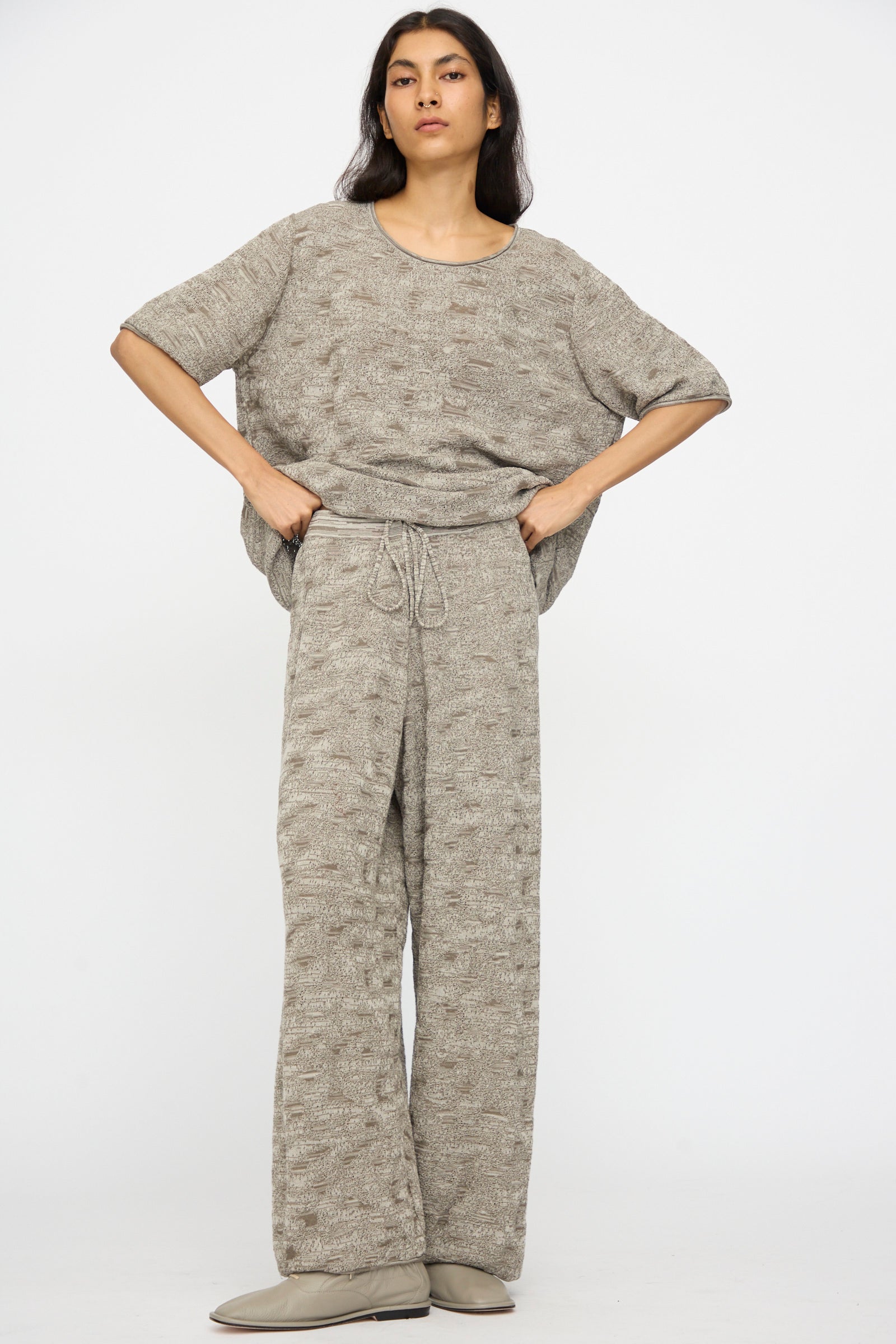 Person standing against a plain background wearing Lauren Manoogian's Terra Pants in Rock Marl, which are relaxed with an elastic waist, paired with a loose-fitting textured beige top and light-colored shoes.