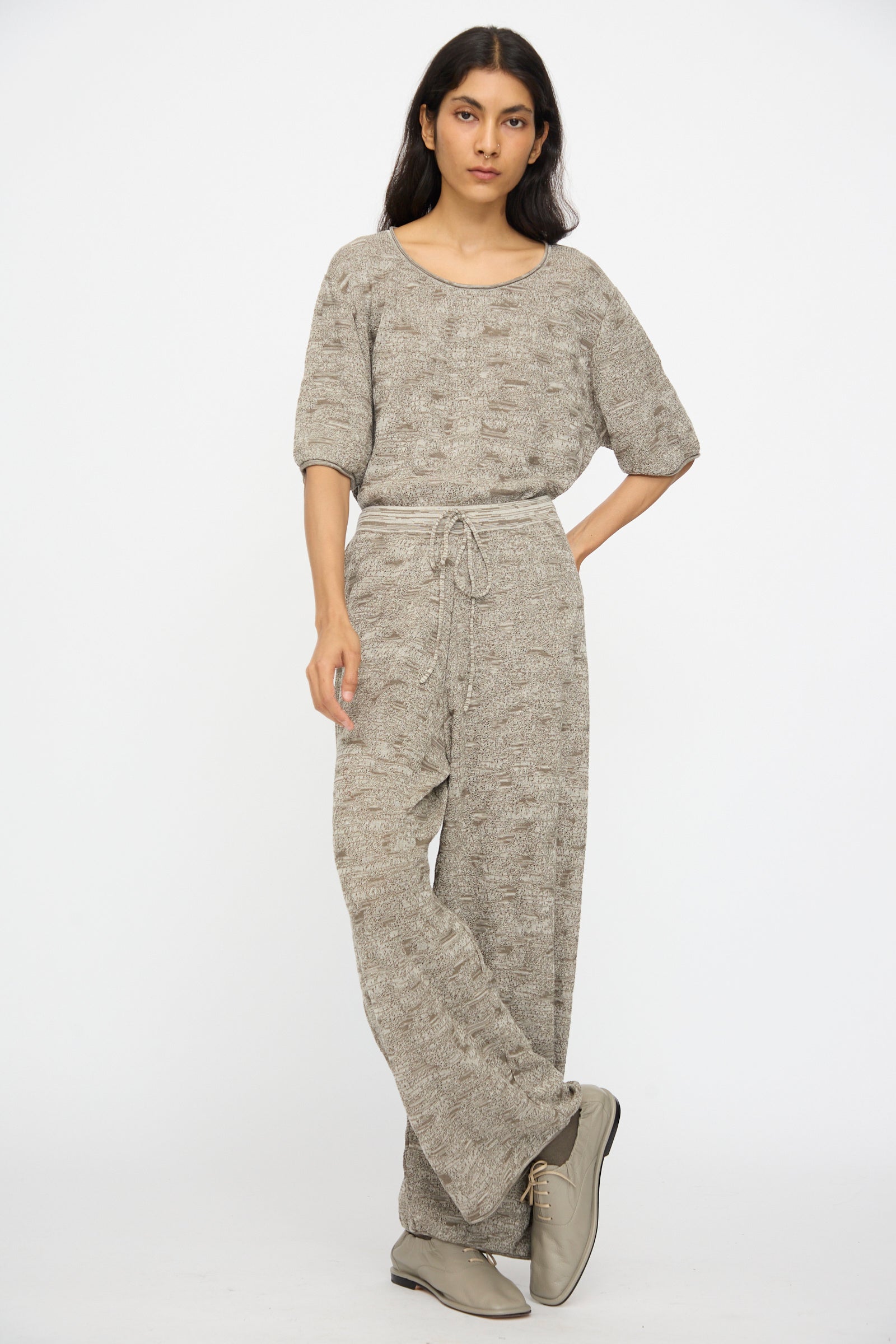 A woman stands against a plain background, wearing Lauren Manoogian’s Terra Pants in Rock Marl—a relaxed, textured grey knit with an elastic waist tie—paired with a matching top and beige lace-up shoes.