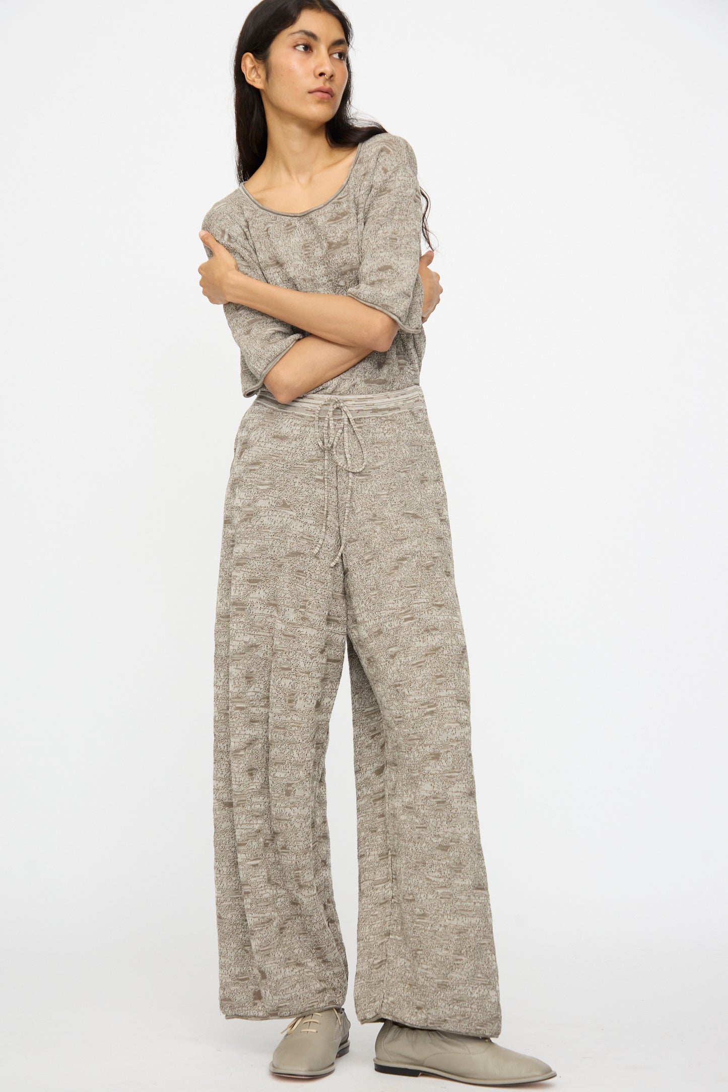 A person stands with arms crossed, wearing Lauren Manoogian’s Terra Pants in Rock Marl—relaxed fit with an elastic waist—paired with a loose light gray knit top and gray shoes, set against a plain white background.