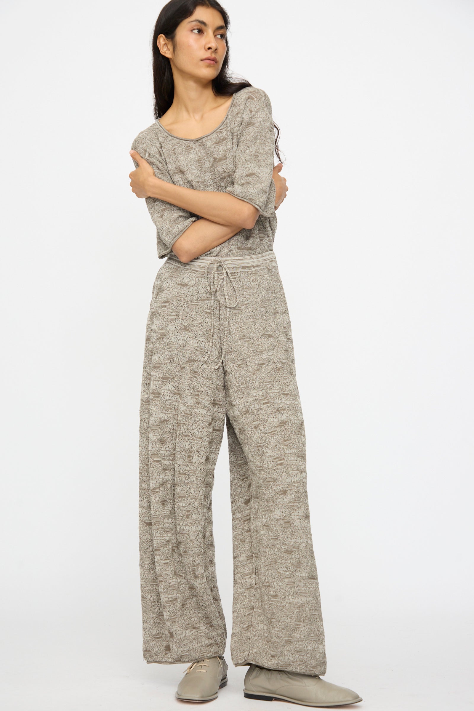 A person stands with arms crossed, wearing Lauren Manoogian’s Terra Pants in Rock Marl—relaxed fit with an elastic waist—paired with a loose light gray knit top and gray shoes, set against a plain white background.