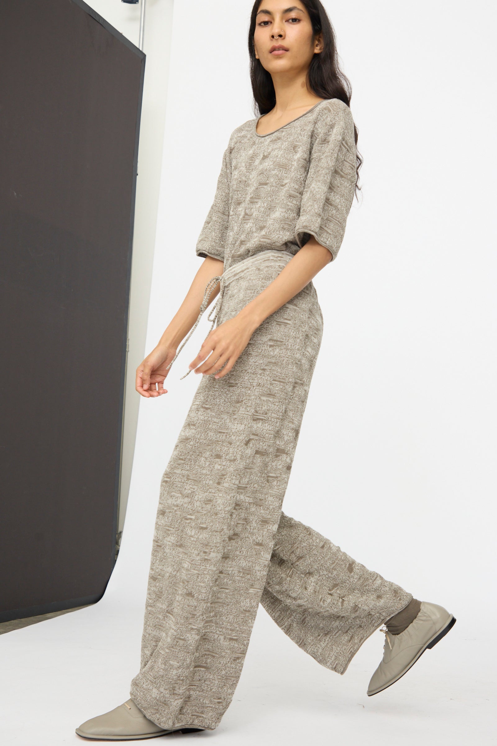 A person in Lauren Manoogian’s Terra Pants in Rock Marl—relaxed pima cotton pants with an elastic waist—paired with a loose beige top and gray shoes, walking in a minimalist studio.