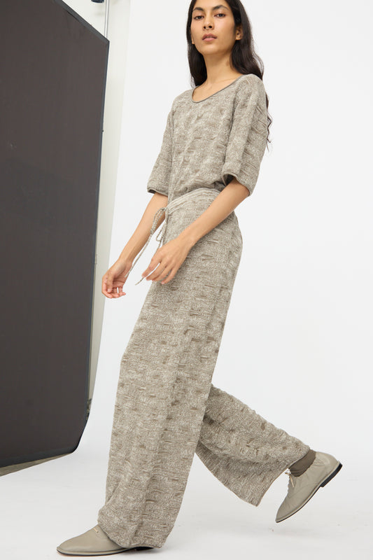 A person in Lauren Manoogian’s Terra Pants in Rock Marl—relaxed pima cotton pants with an elastic waist—paired with a loose beige top and gray shoes, walking in a minimalist studio.