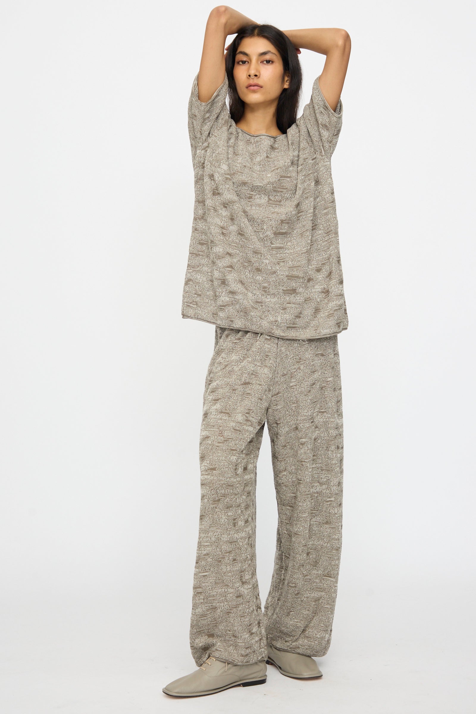 A person stands with arms raised, wearing the Lauren Manoogian Terra Tee in Rock Marl and matching wide-leg pants, paired with light-colored shoes against a plain white background.