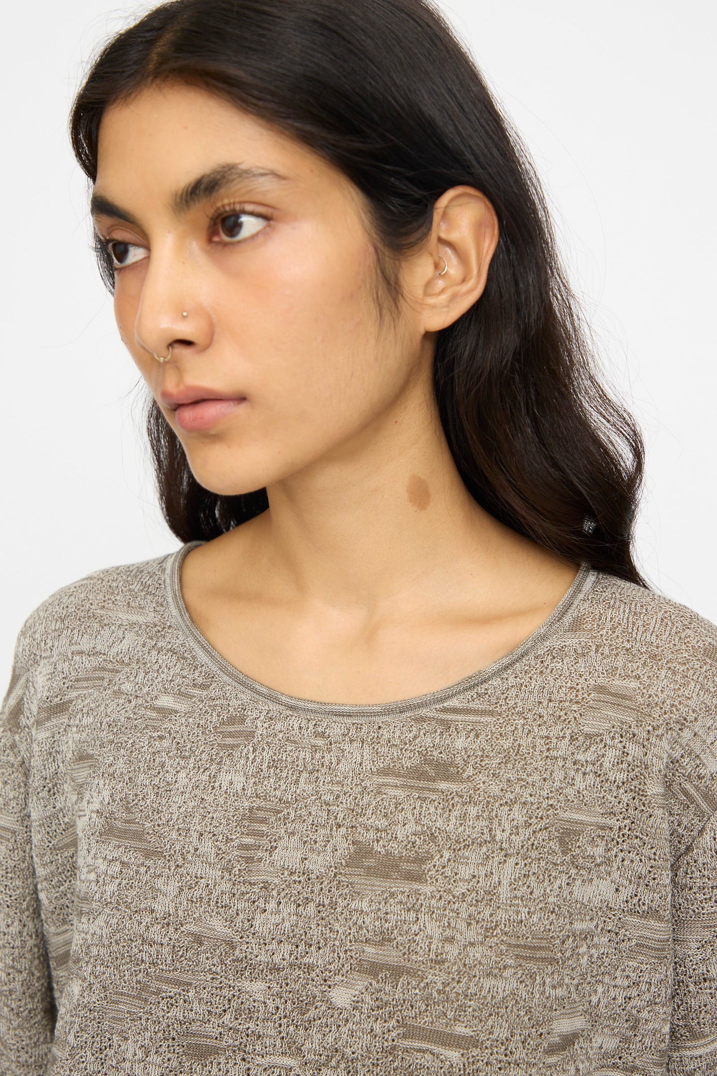 A person with long dark hair and a nose ring, wearing the Lauren Manoogian Terra Tee in Rock Marl, looks to the side. A brown birthmark is visible on their neck.