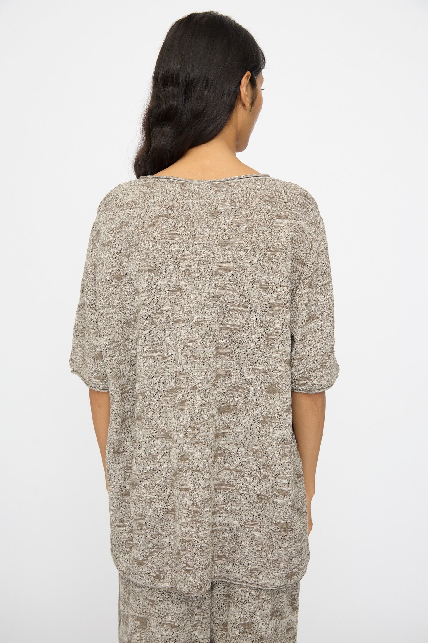 A person with long dark hair stands facing away, wearing the Lauren Manoogian Terra Tee in Rock Marl—a loose-fitting, patterned beige and gray top crafted from soft mercerized pima cotton—against a plain white background.