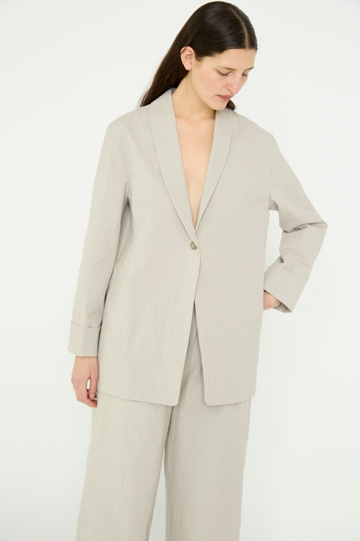 A person wearing Lauren Manoogian's Texture Blazer in Pewter, featuring light crinkled cotton and an oversized fit, pairs it with wide-leg pants while standing against a plain white background, looking downwards.