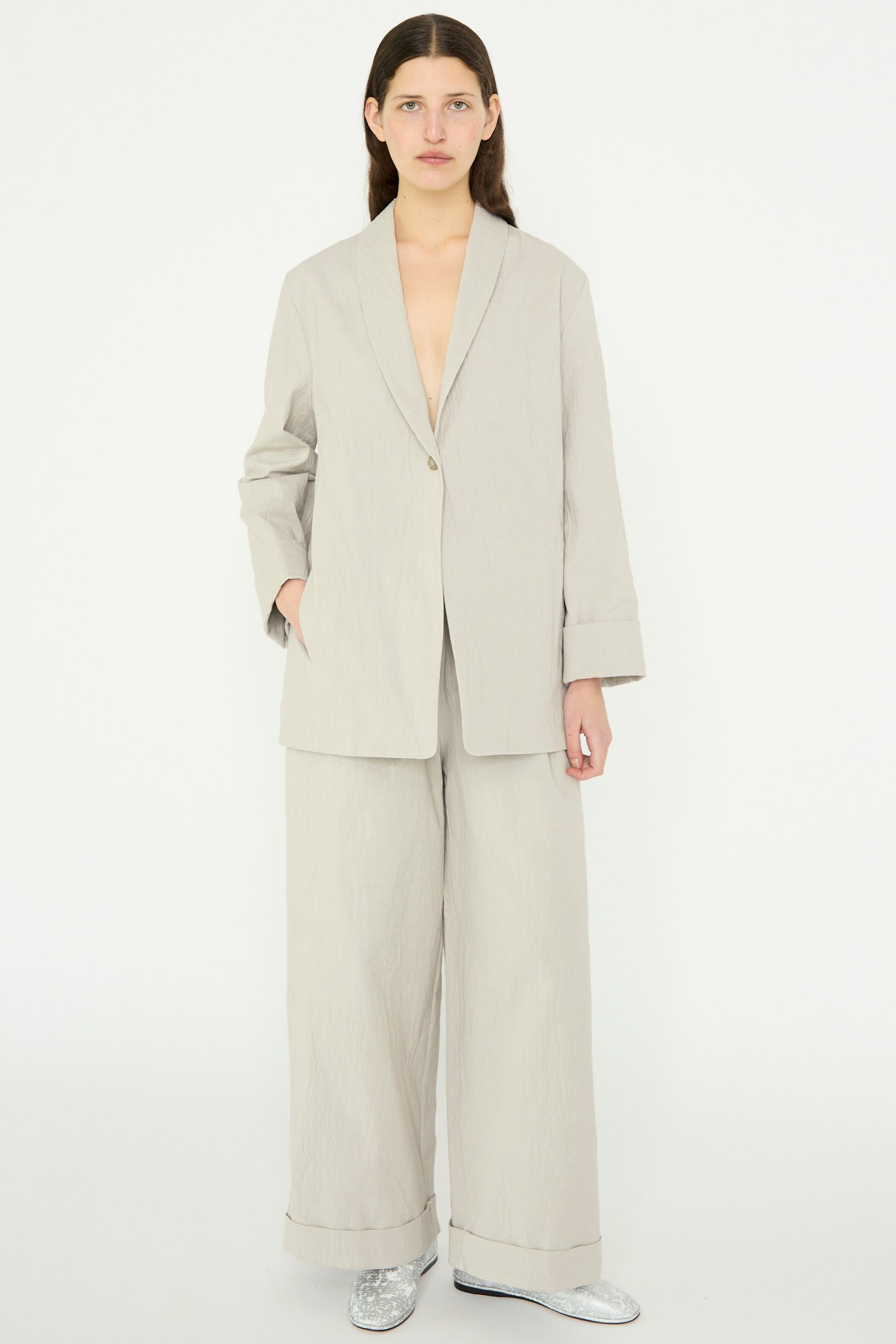 A model stands against a plain background in a Lauren Manoogian Texture Blazer in Pewter, paired with wide-legged pants and silver shoes. The blazer features an oversized design with a single-button closure, and the person keeps one hand in their pocket.