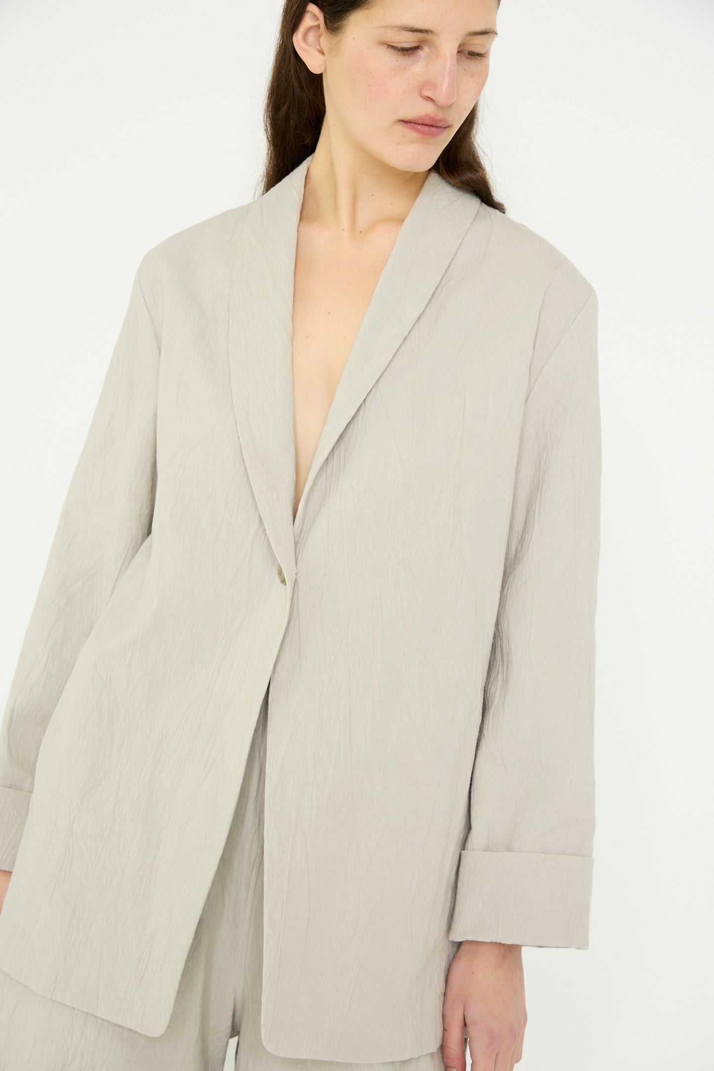 A person stands against a plain white background, wearing the Lauren Manoogian Texture Blazer in Pewter and matching pants The outfit features a loose fit and oversized design with a shawl collar.