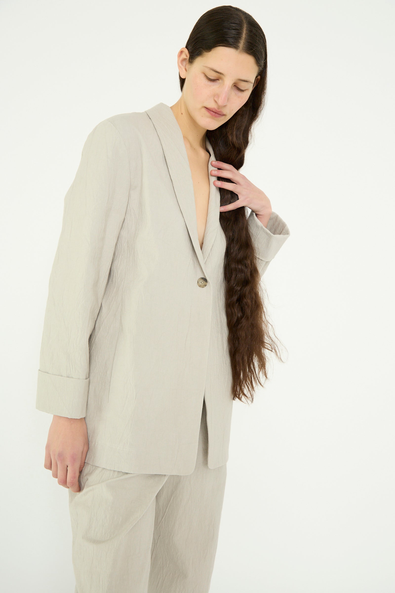 A long-haired person in Lauren Manoogian's oversized Texture Blazer in Pewter touches their hair and looks down 
against a plain white background.