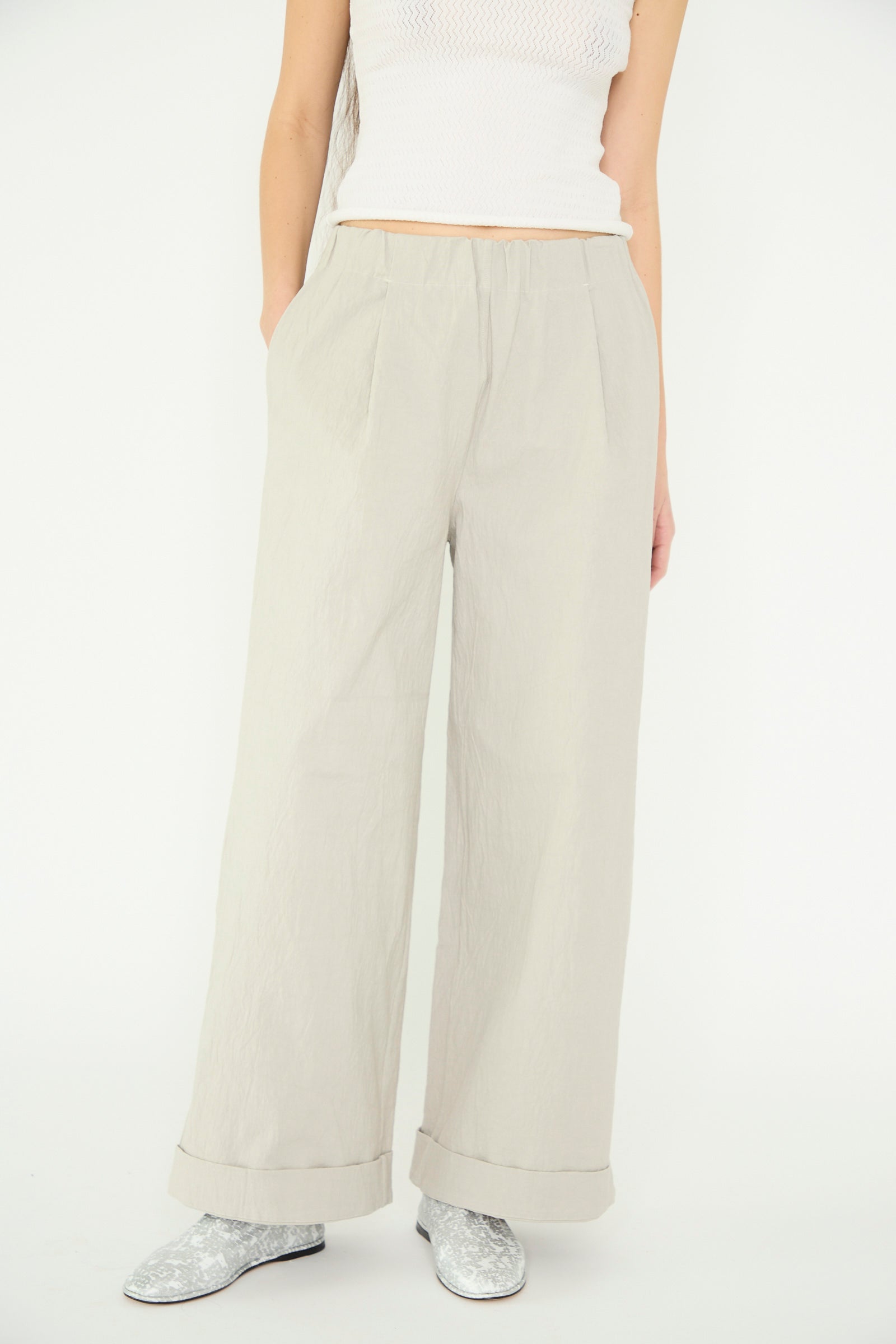 A person wearing a white sleeveless top and Lauren Manoogian's Texture Trouser in Pewter, featuring high-waist wide-leg design in crinkled cotton blend, with hands in pockets, paired with silver shoes against a plain white background.