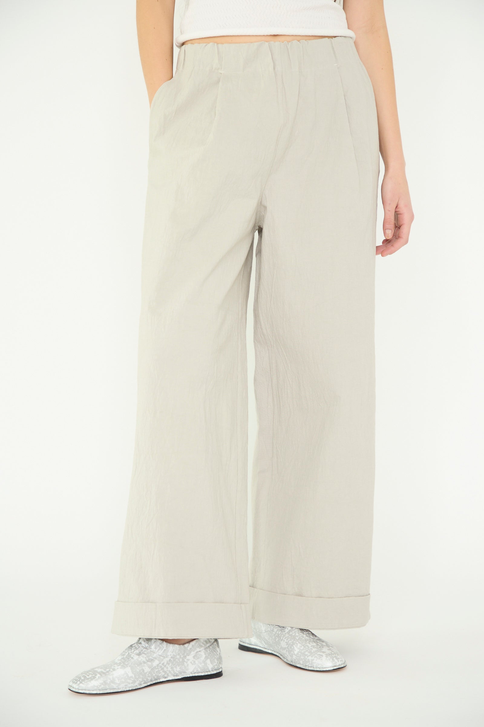 Wearing the Texture Trouser in pewter by Lauren Manoogian, a person stands against a plain backdrop, with silver shoes, one hand in a pocket. 