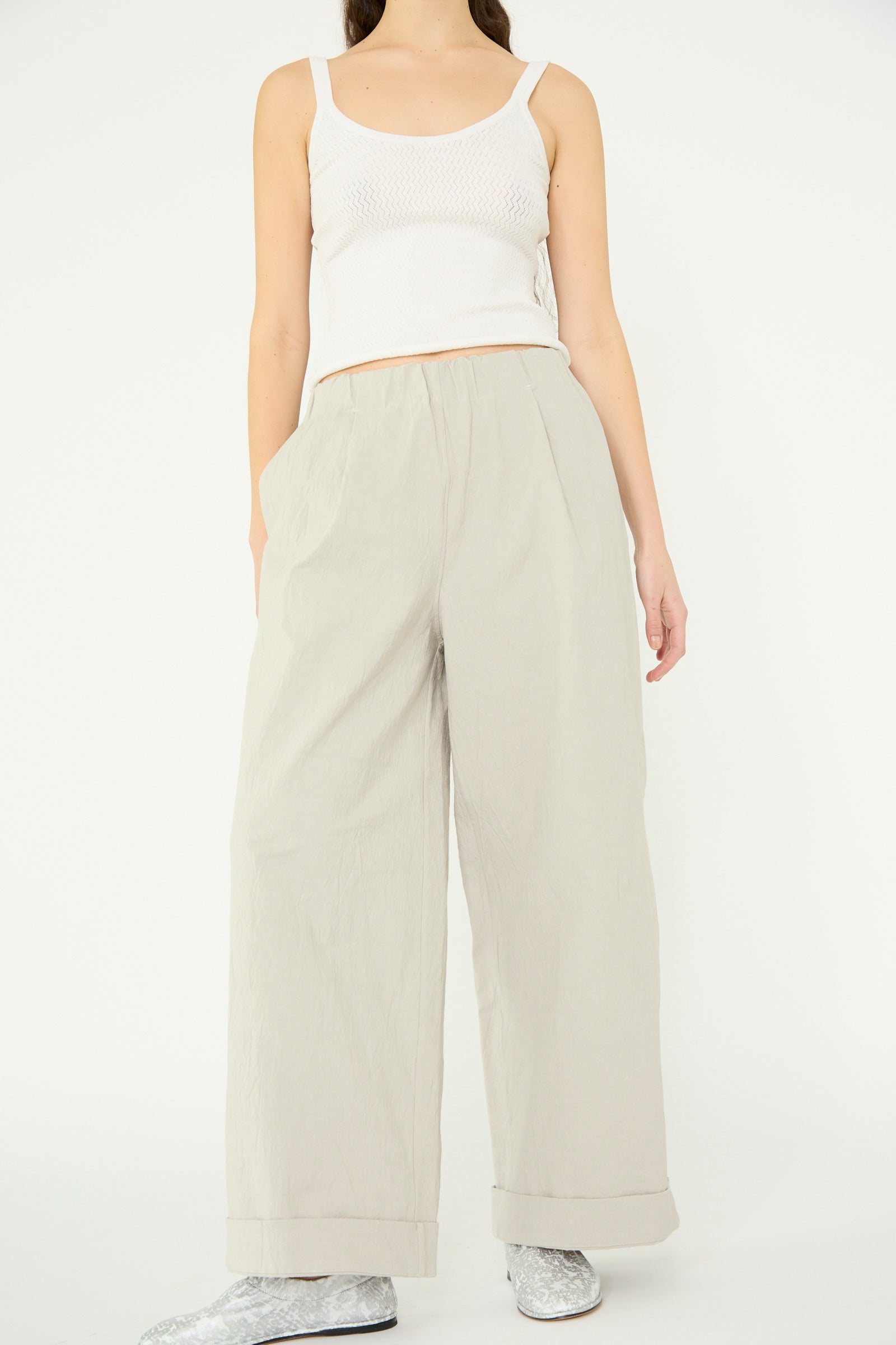 A person stands against a plain background wearing a white sleeveless top and Lauren Manoogian's Texture Trouser in Pewter, light gray high-waist wide-leg pants made from crinkled cotton blend fabric, complemented by silver shoes.
