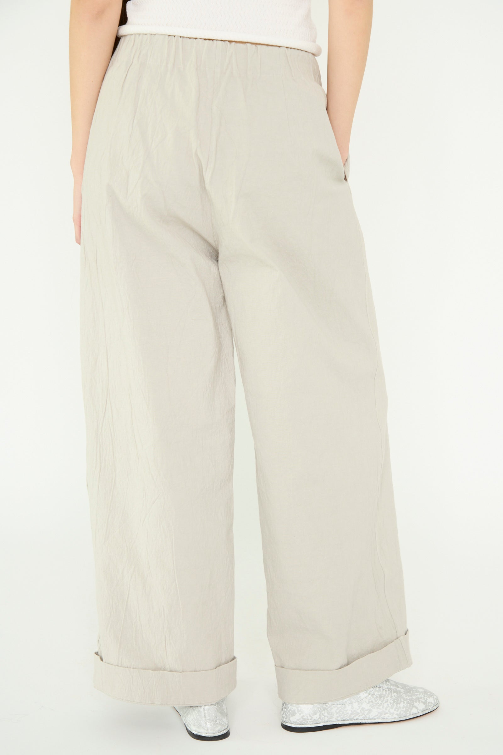 A person wears Lauren Manoogian's Texture Trouser in Pewter, high-waist wide-leg pants made from a crinkled cotton blend, paired with silver shoes, standing with their back to the camera.