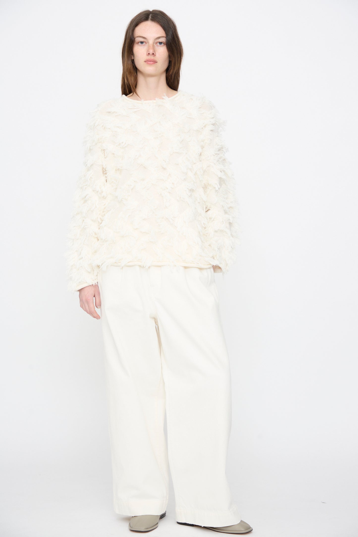A person stands against a plain white background, wearing the Lauren Manoogian Tufted Pullover in Bone, paired with wide-leg white pants and beige shoes.