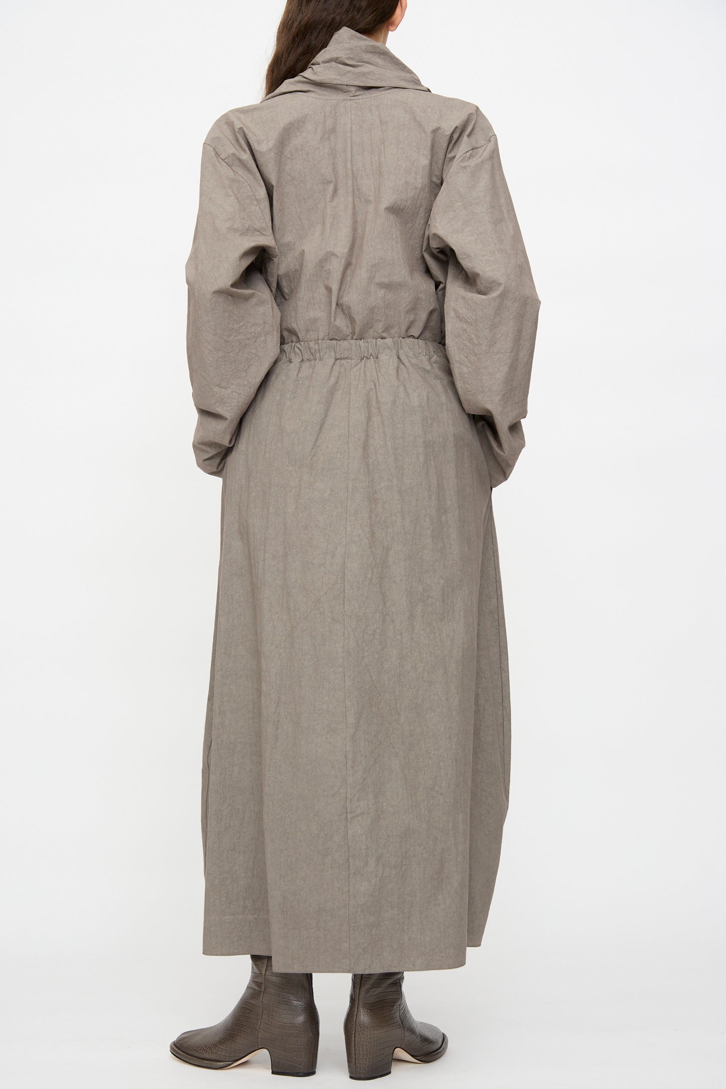 A person stands with their back to the camera, wearing a long gray coat with an elastic waistband; the Lauren Manoogian Twist Skirt in Cinder is visible above brown heeled boots.