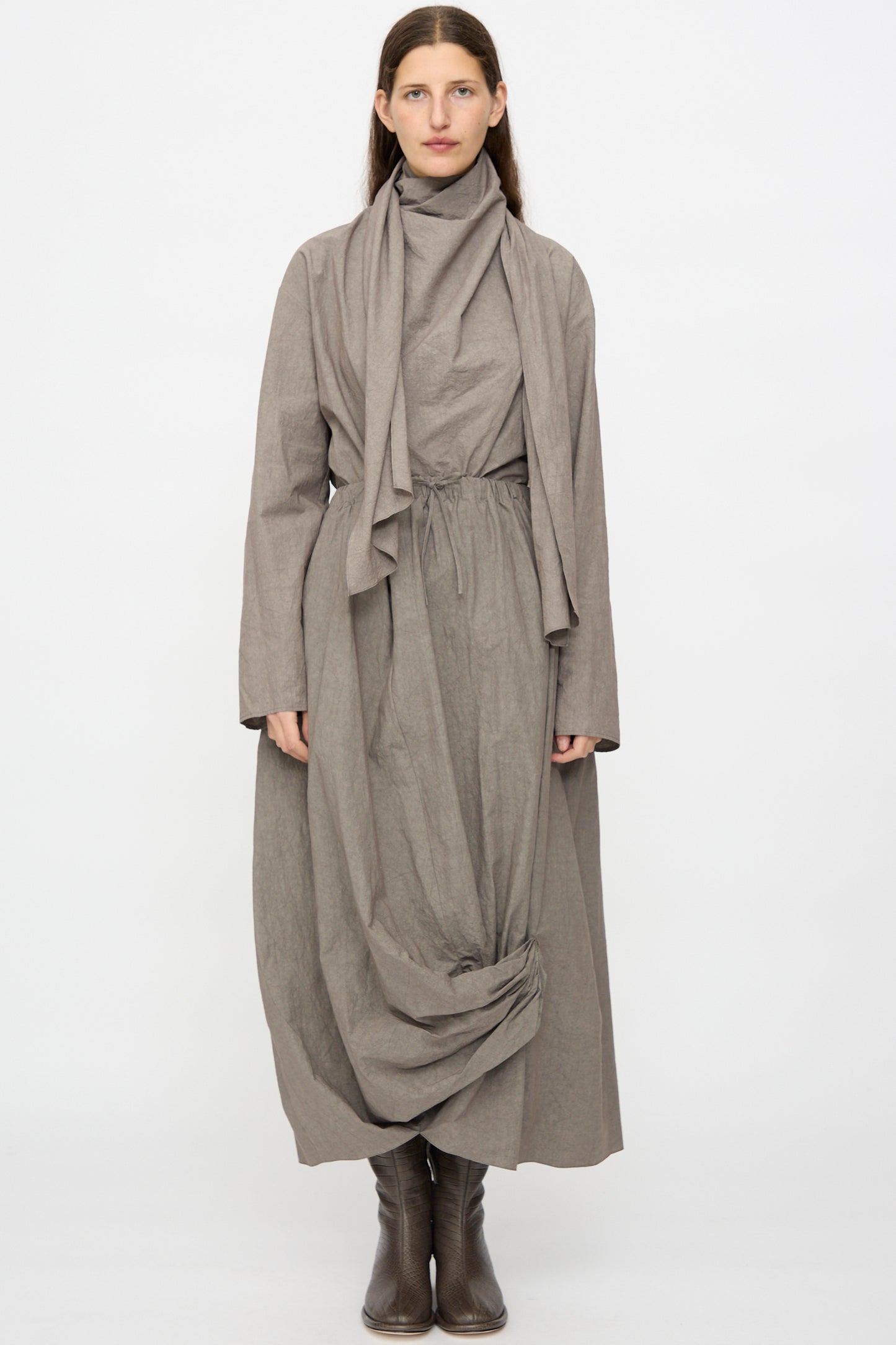 A woman stands against a plain white background wearing Lauren Manoogian's Twist Skirt in Cinder, paired with a loose long-sleeved taupe dress featuring a high collar, scarf, and gathered hem detail, styled with brown boots.