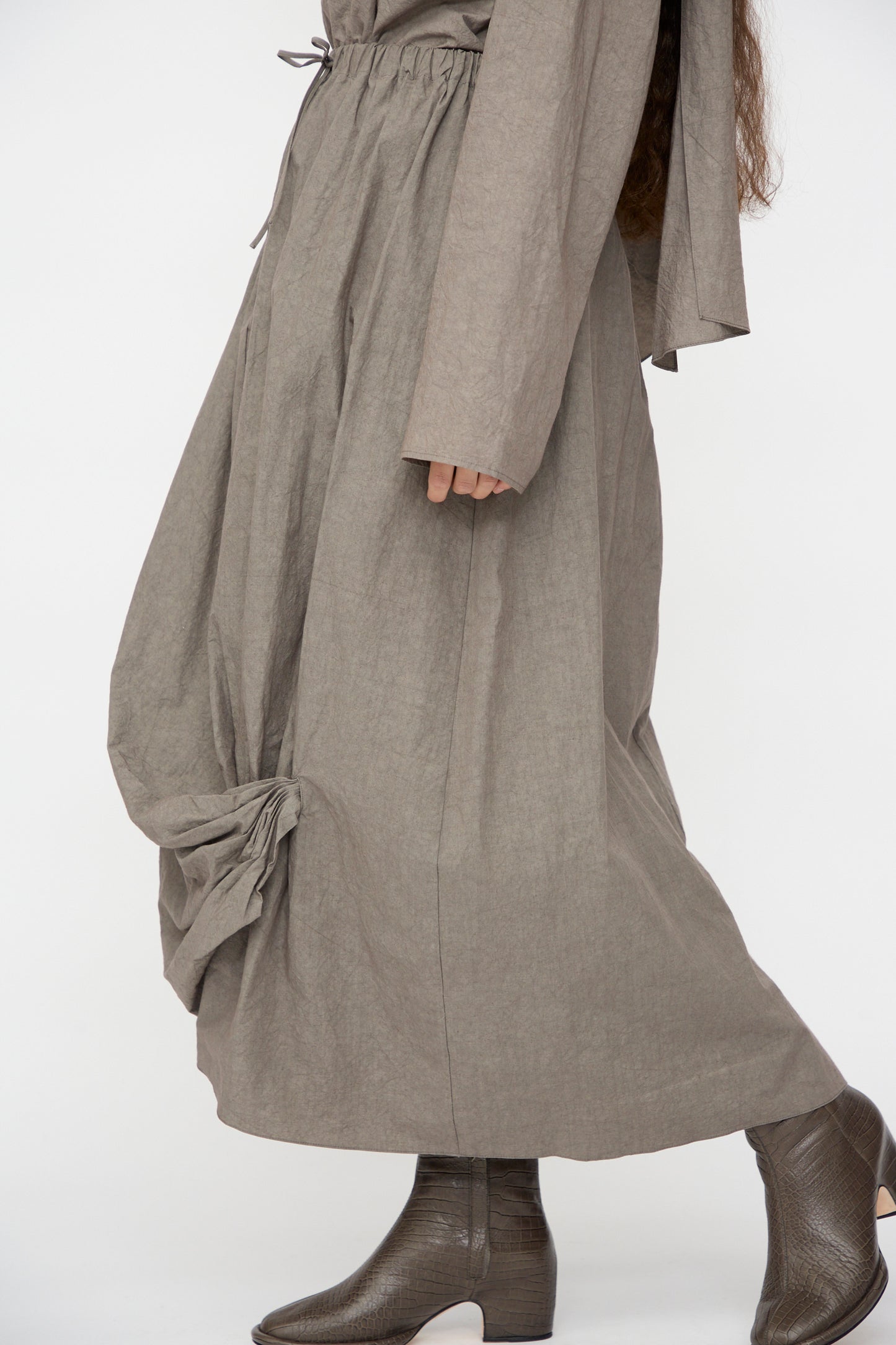 Model wears the Lauren Manoogian Twist Skirt in Cinder, a long, loose taupe cotton-linen skirt with gathered hem details, paired with textured ankle boots. Photographed against a plain white background.