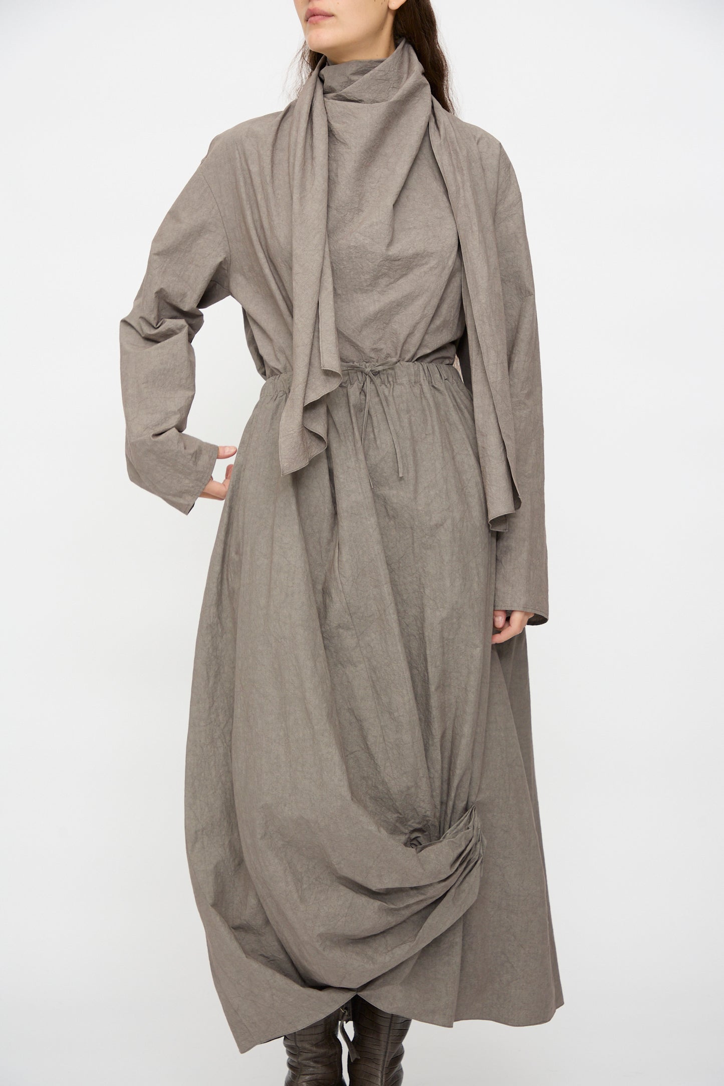 A person wearing the Lauren Manoogian Twist Skirt in Cinder—a loose, long taupe cotton-linen skirt with a high neck, draped scarf detail, cinched waist, and gathered hem—artfully crafted in Japan.