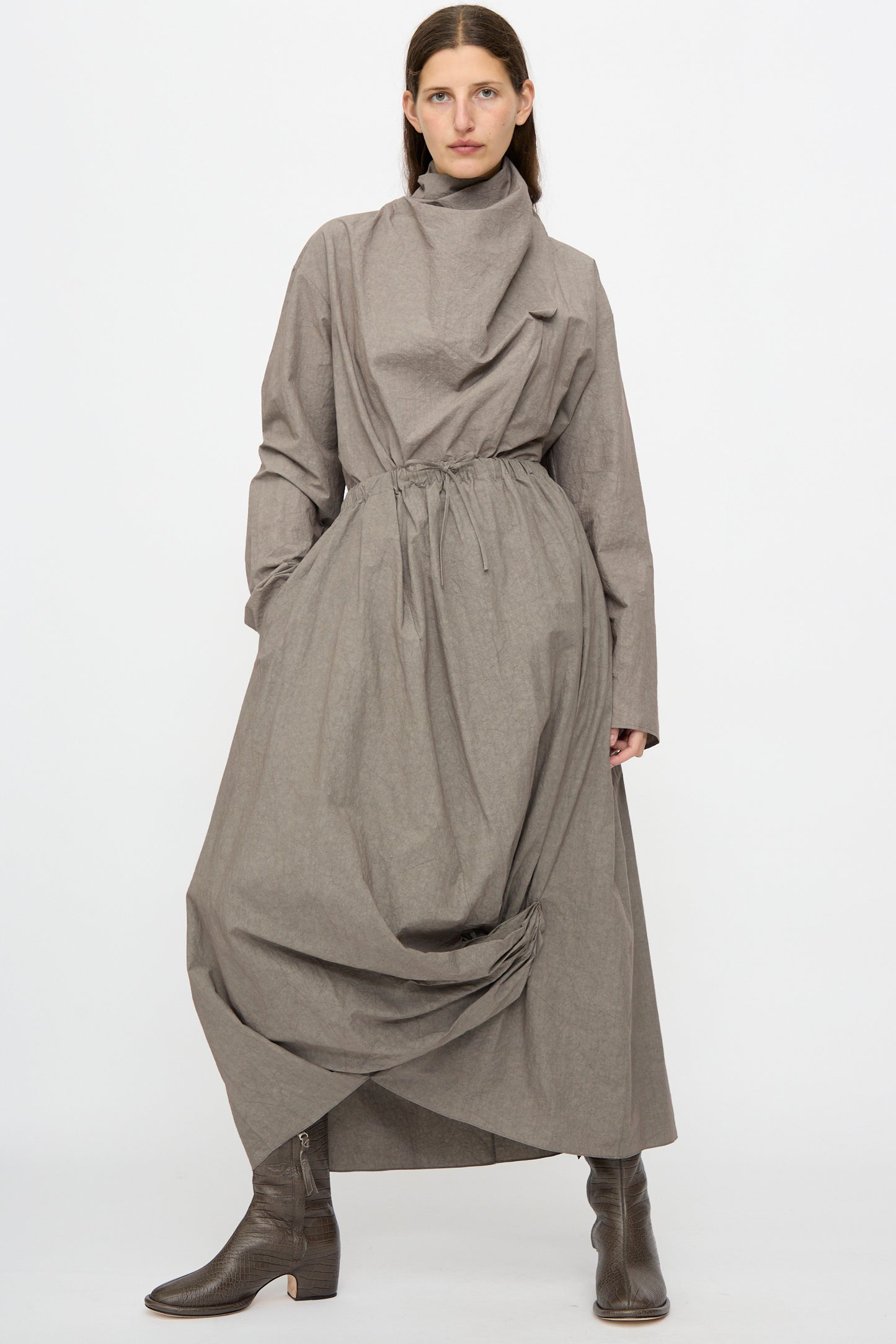 A person models the Lauren Manoogian Twist Skirt in Cinder, styled with heeled brown boots and a loose gray top, standing against a plain white background.