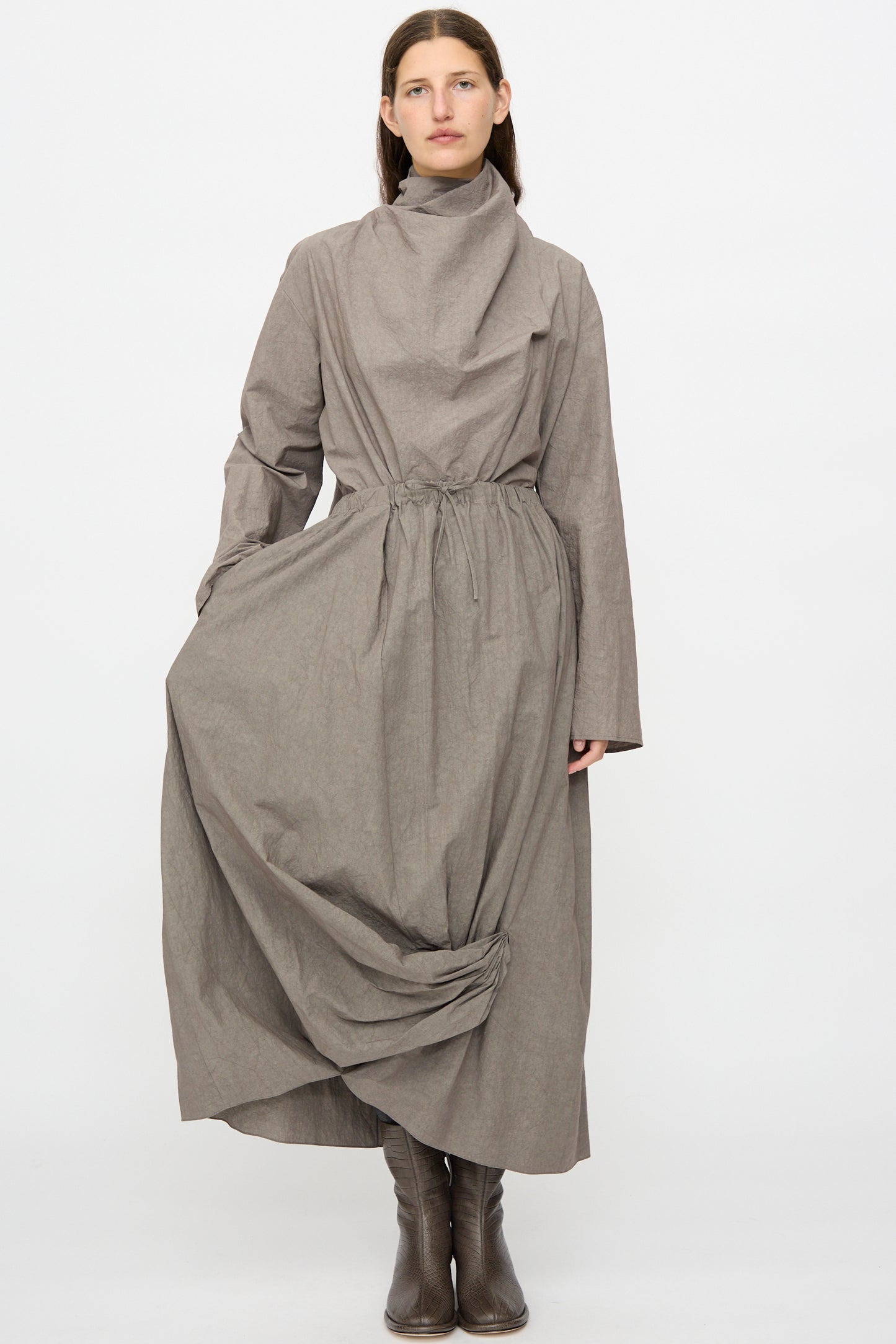 A person stands against a plain background wearing the Lauren Manoogian Twist Skirt in Cinder—a long, loose-fitting cotton-linen skirt with a high neck, gathered waist, and draped silhouette—paired with brown boots. Thoughtfully made in Japan.