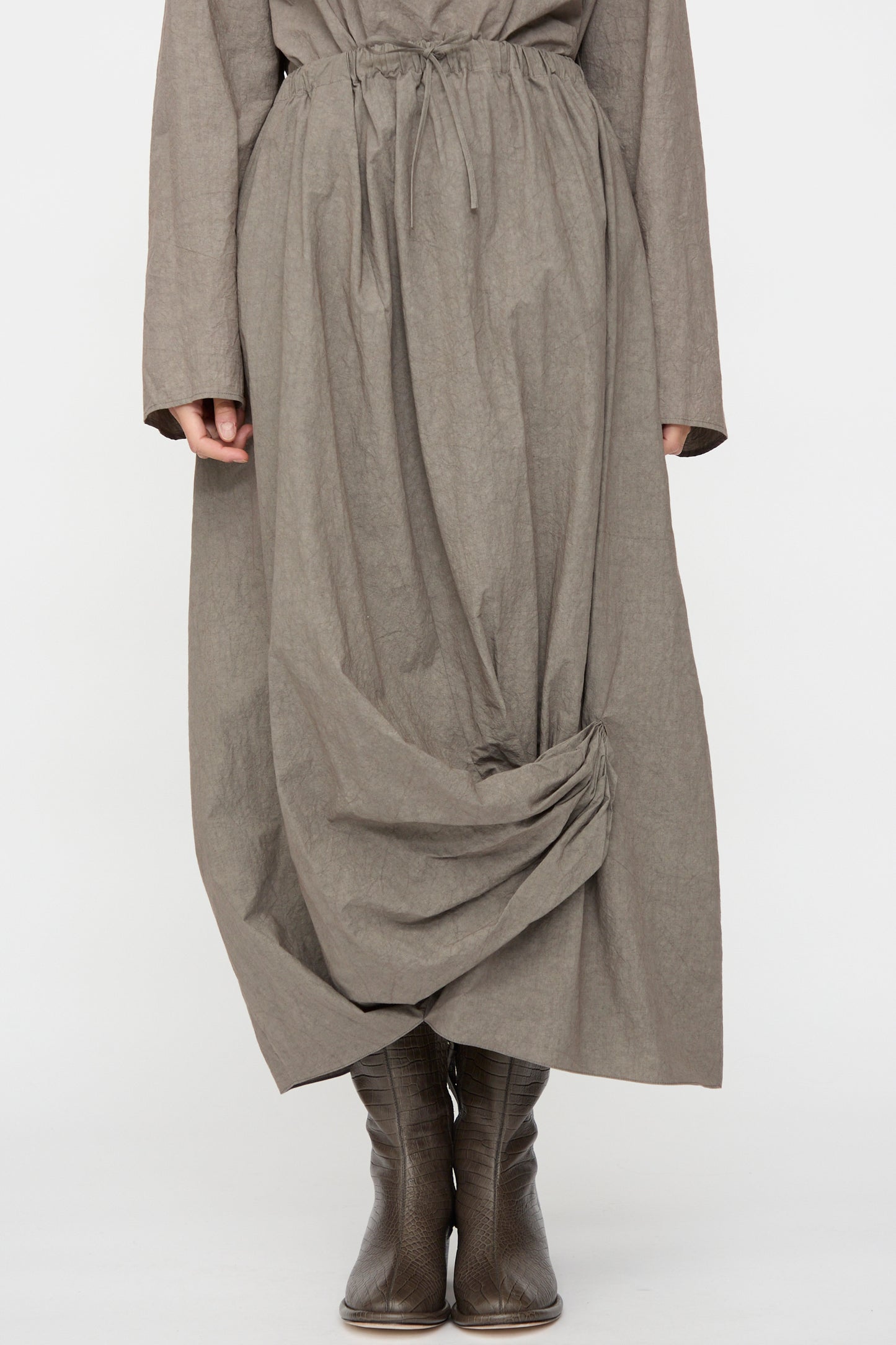 A person wearing the Lauren Manoogian Twist Skirt in Cinder, a long, loose-fitting cotton linen piece with gathered hem detail, paired with brown leather boots. Only the lower half of this Japan-made outfit is visible.