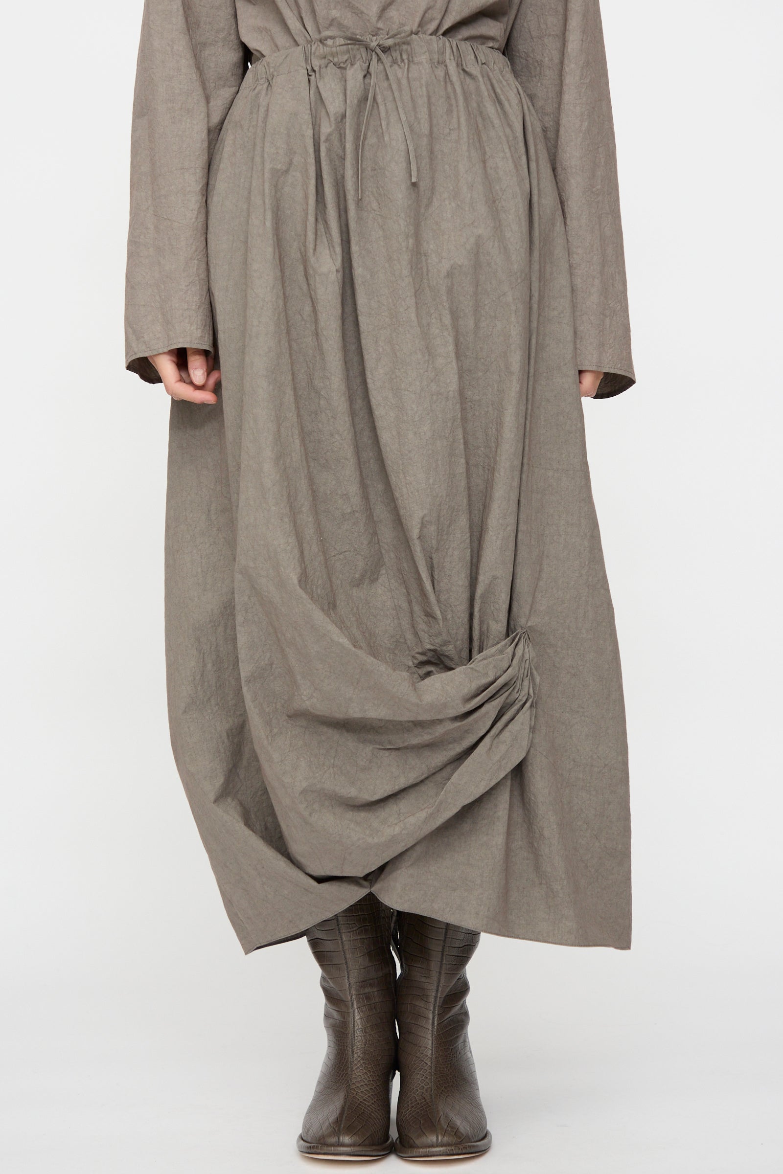 A person wearing the Lauren Manoogian Twist Skirt in Cinder, a long, loose-fitting cotton linen piece with gathered hem detail, paired with brown leather boots. Only the lower half of this Japan-made outfit is visible.