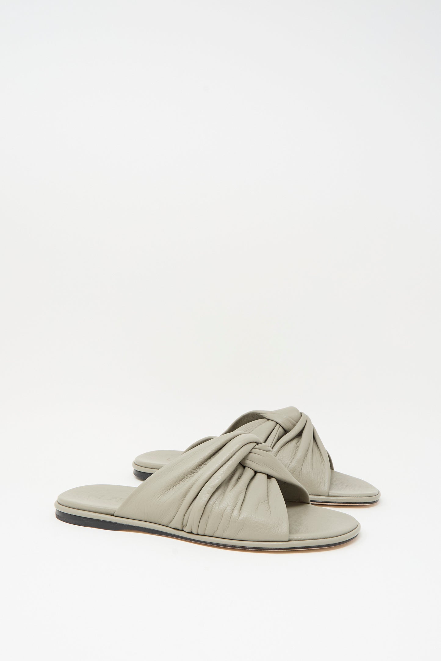The Lauren Manoogian Twist Slide in Stone features light gray leather with twisted, ruched straps and flat soles, handmade and showcased against a plain white background.