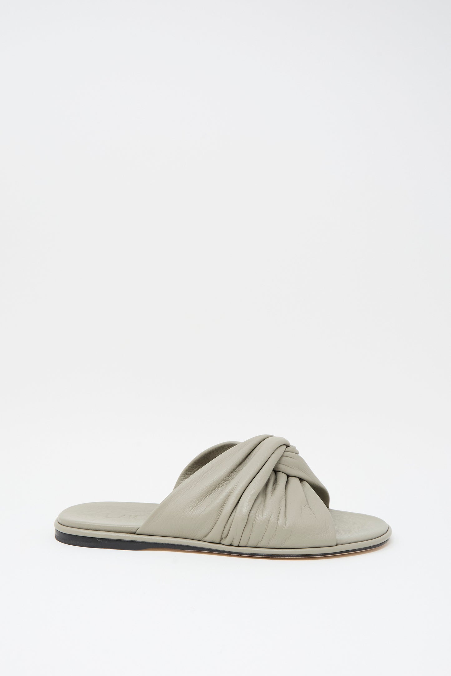 The Lauren Manoogian Twist Slide in Stone is a flat, light gray sandal featuring a gathered twist leather strap and a slightly raised black sole, displayed on a plain white background.