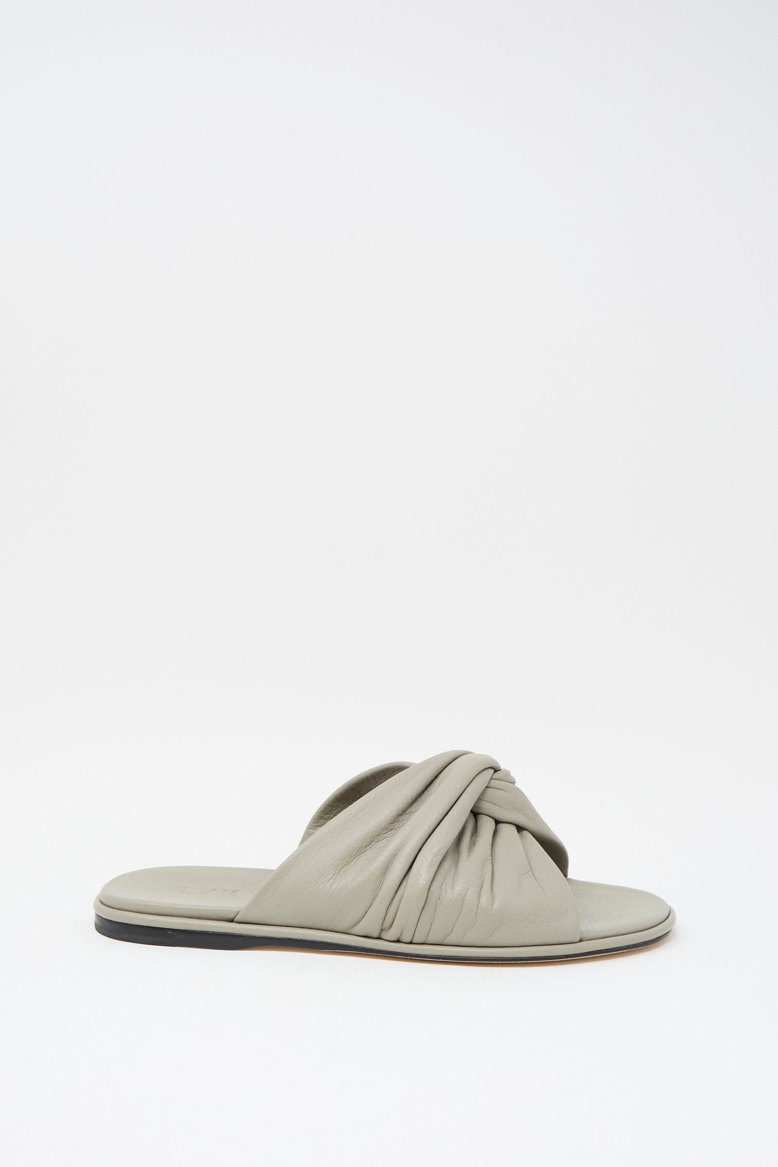 The Lauren Manoogian Twist Slide in Stone is a flat, light gray sandal featuring a gathered twist leather strap and a slightly raised black sole, displayed on a plain white background.